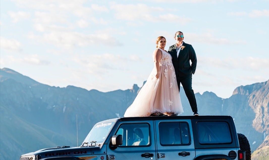A newlywed couple stands atop of their Jeep overlooking a mountain. One is in a dress, one is in a suit.