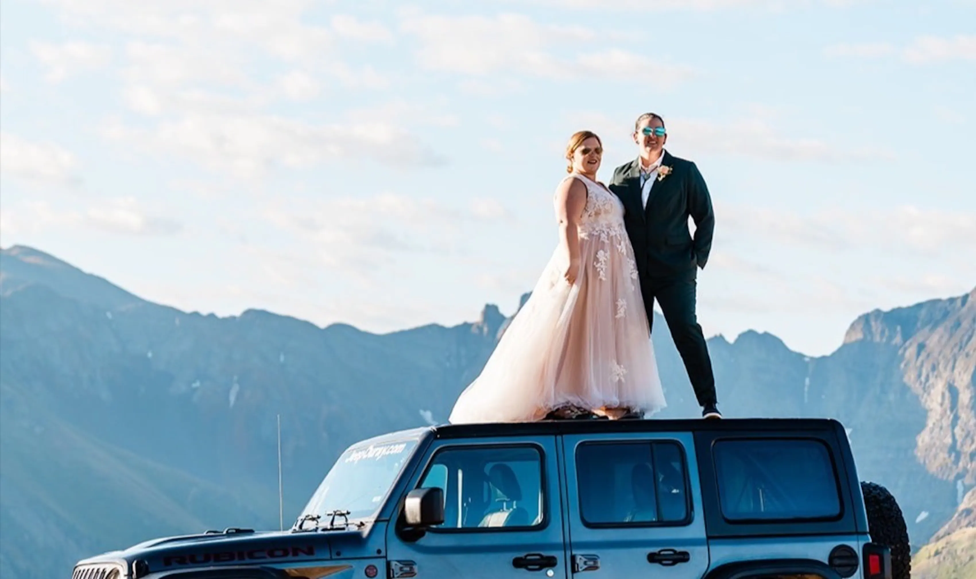 A newlywed couple stands atop of their Jeep overlooking a mountain. One is in a dress, one is in a suit.