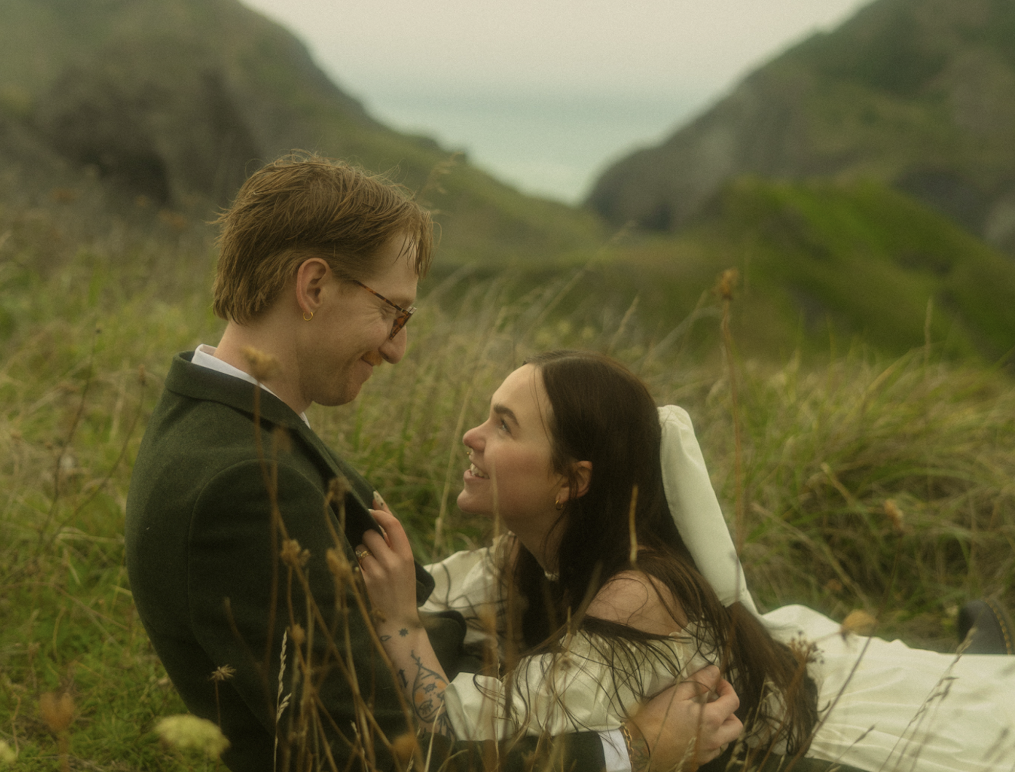 Image of a couple in a secluded field in the mountains laying down, staring at eachother. The woman is in a dress and a small veil on top of the man, who is in a suit.