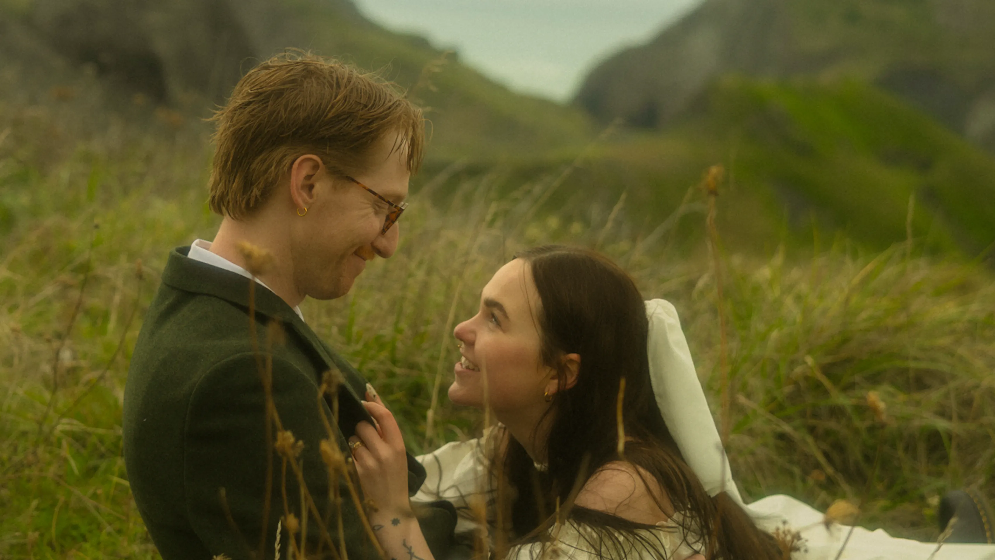 Image of a couple in a secluded field in the mountains laying down, staring at eachother. The woman is in a dress and a small veil on top of the man, who is in a suit.