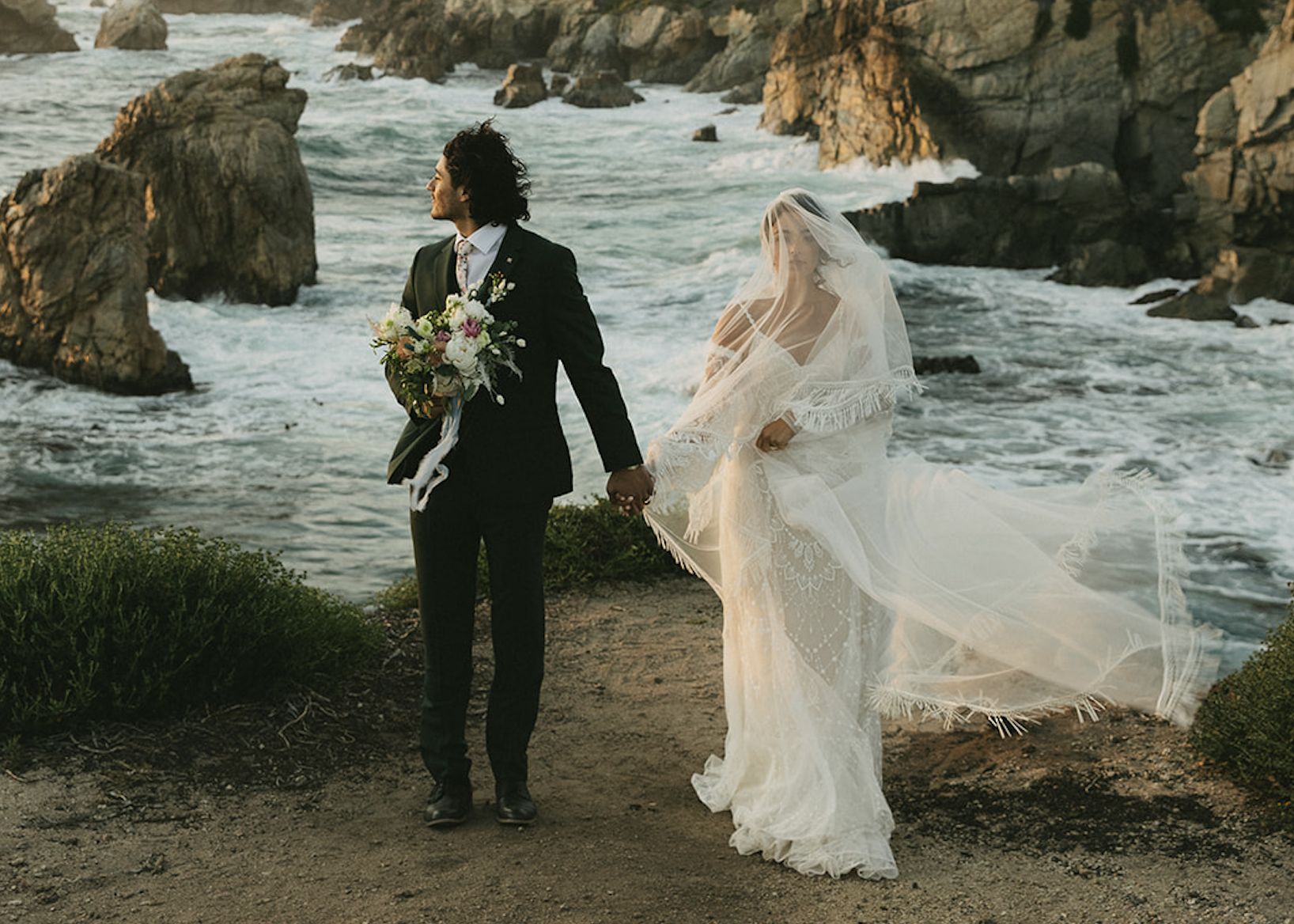 An image of a couple holding hands by the shore at a beach. She is in her wedding gown as her veil is swept by the wind. He looks to the side in his suit, holding a bouquet in his free hand.