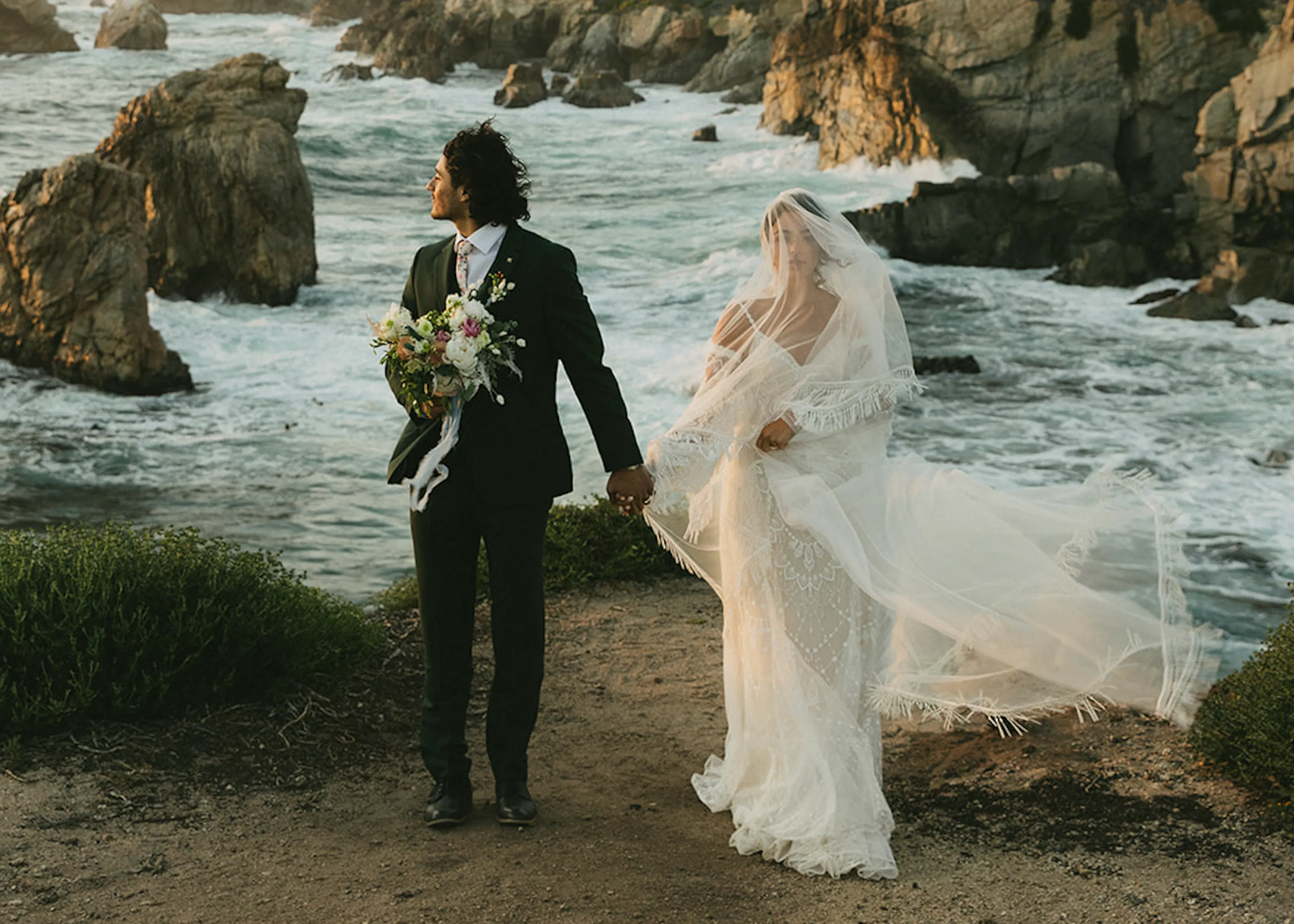 An image of a couple holding hands by the shore at a beach. She is in her wedding gown as her veil is swept by the wind. He looks to the side in his suit, holding a bouquet in his free hand.