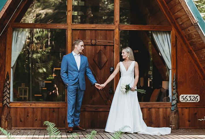 A man and woman stand in front of their cabin rental, holding hands and looking at one another. She is in a white dress holding flowers in her free hand, and he is in a light blue suit.