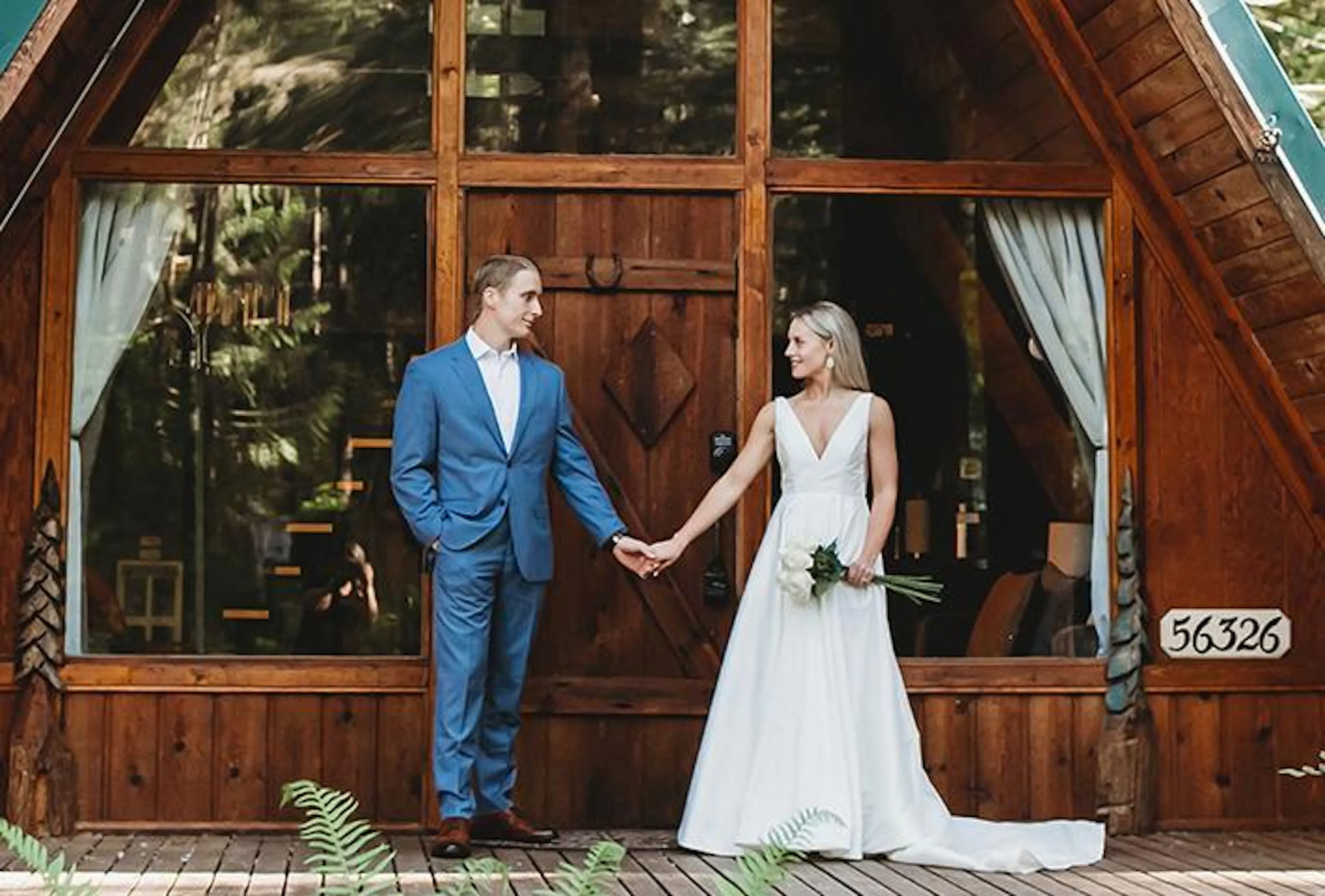 A man and woman stand in front of their cabin rental, holding hands and looking at one another. She is in a white dress holding flowers in her free hand, and he is in a light blue suit.