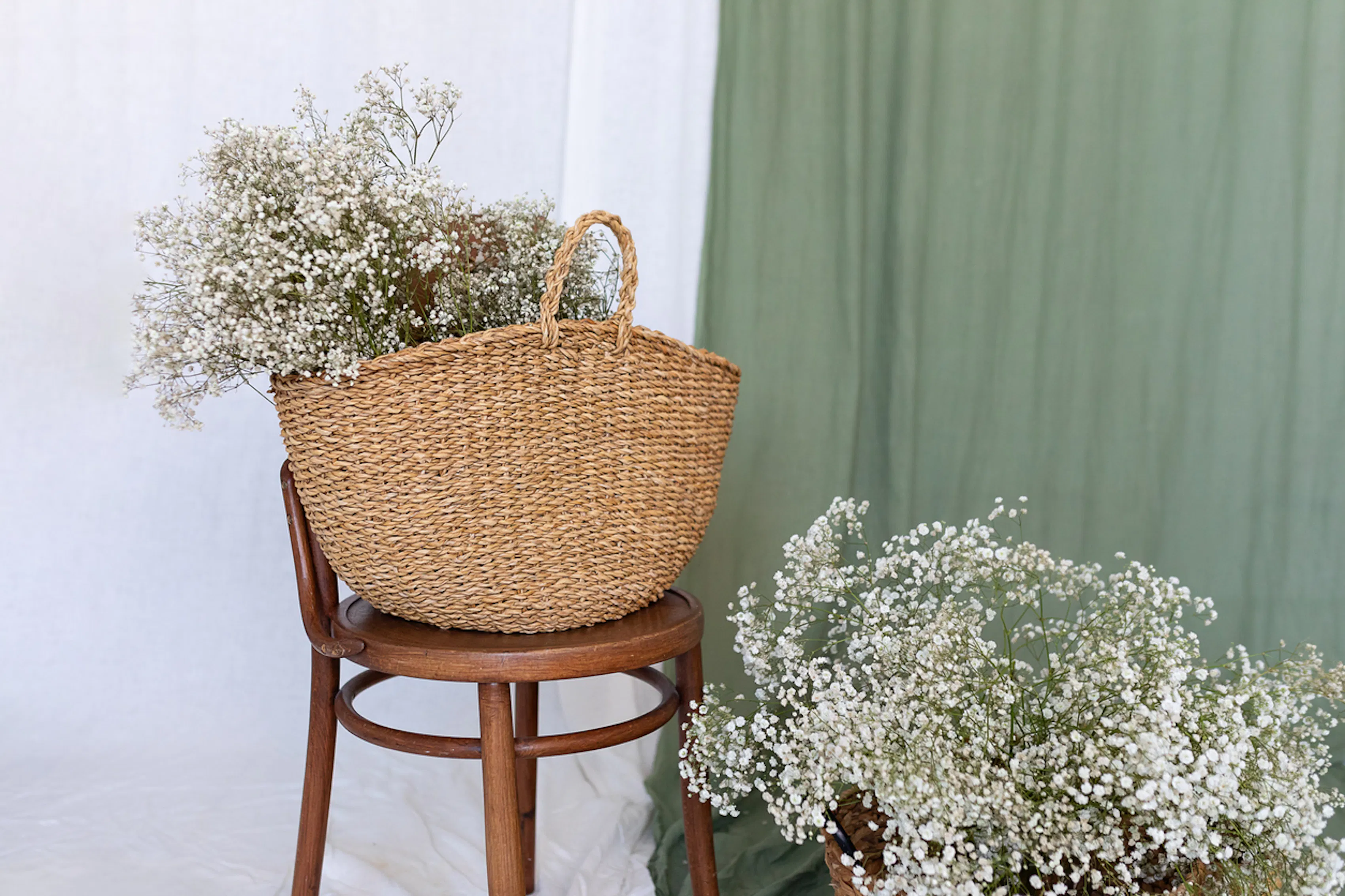 A bouquet of babys breath sits in a basket on a stool in front of a green curtain. There is another basket of the same thing below it.