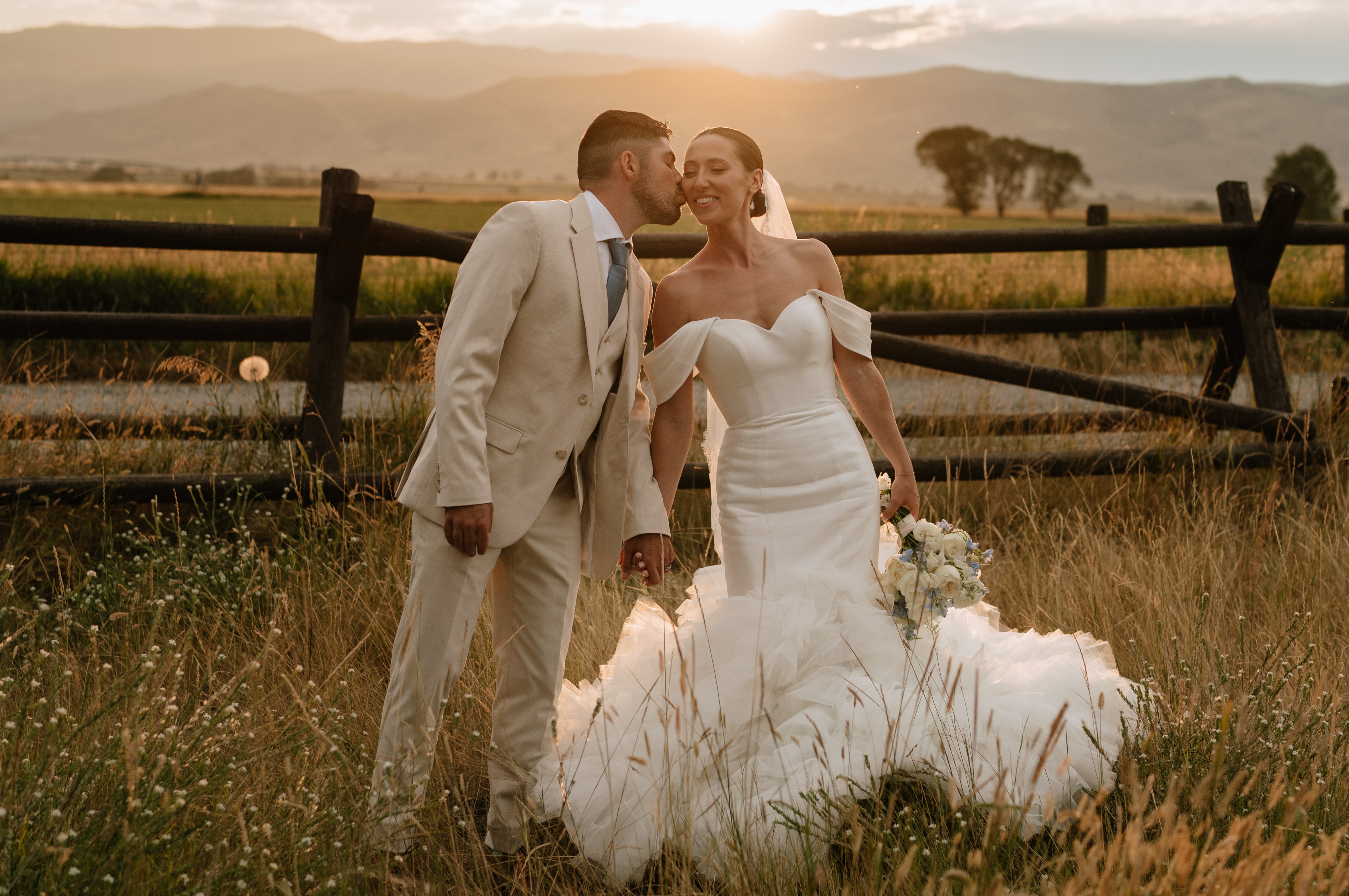 A newlywed husband and wife stand in a field in front of a fence as the sun begins to set. He kisses her on the cheek,