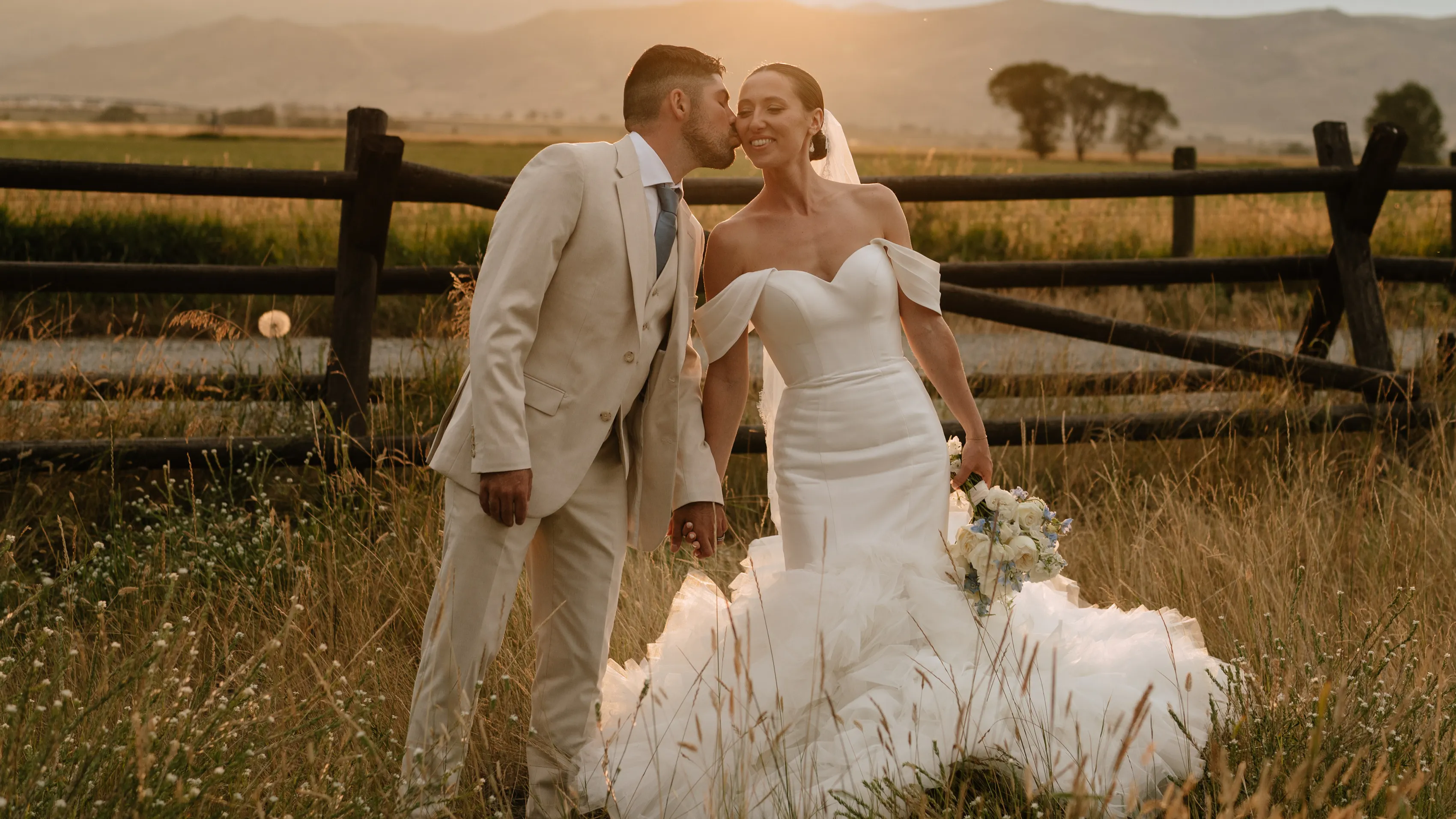 A newlywed husband and wife stand in a field in front of a fence as the sun begins to set. He kisses her on the cheek,