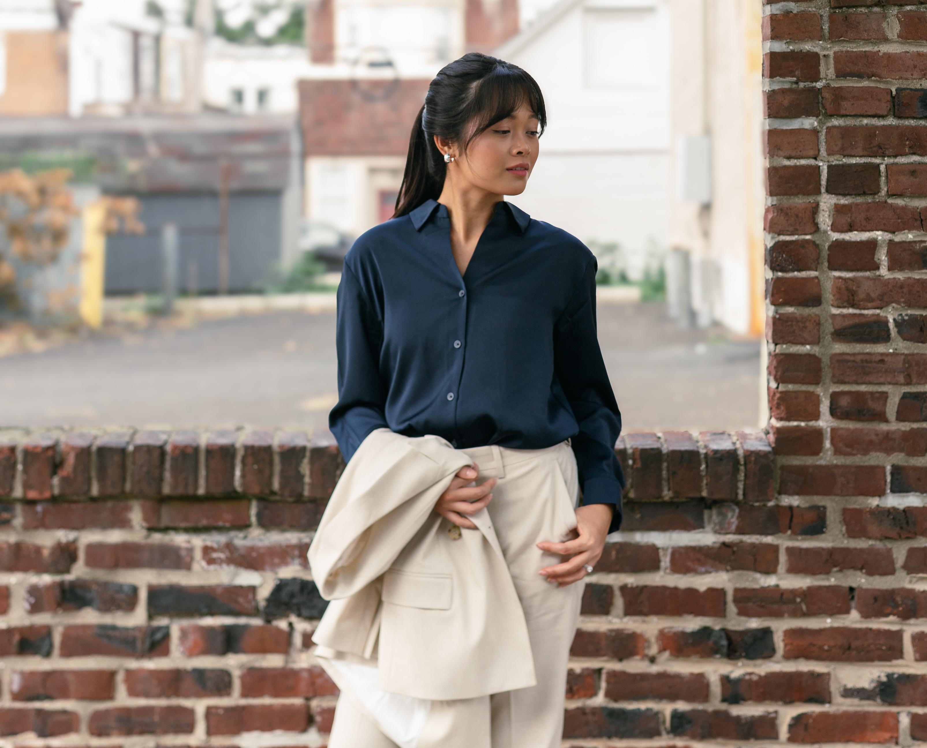 A woman in dressy casual holds her blazer and stands in front of a brick ledge.