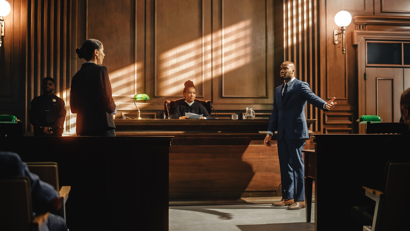 A courtroom scene with a judge sitting at the bench, listening to a lawyer presenting a case. A man in a blue suit stands before the judge, speaking while another person, likely a lawyer, faces him. A security officer stands near the back of the courtroom. The lighting creates dramatic shadows on the wood-paneled walls.