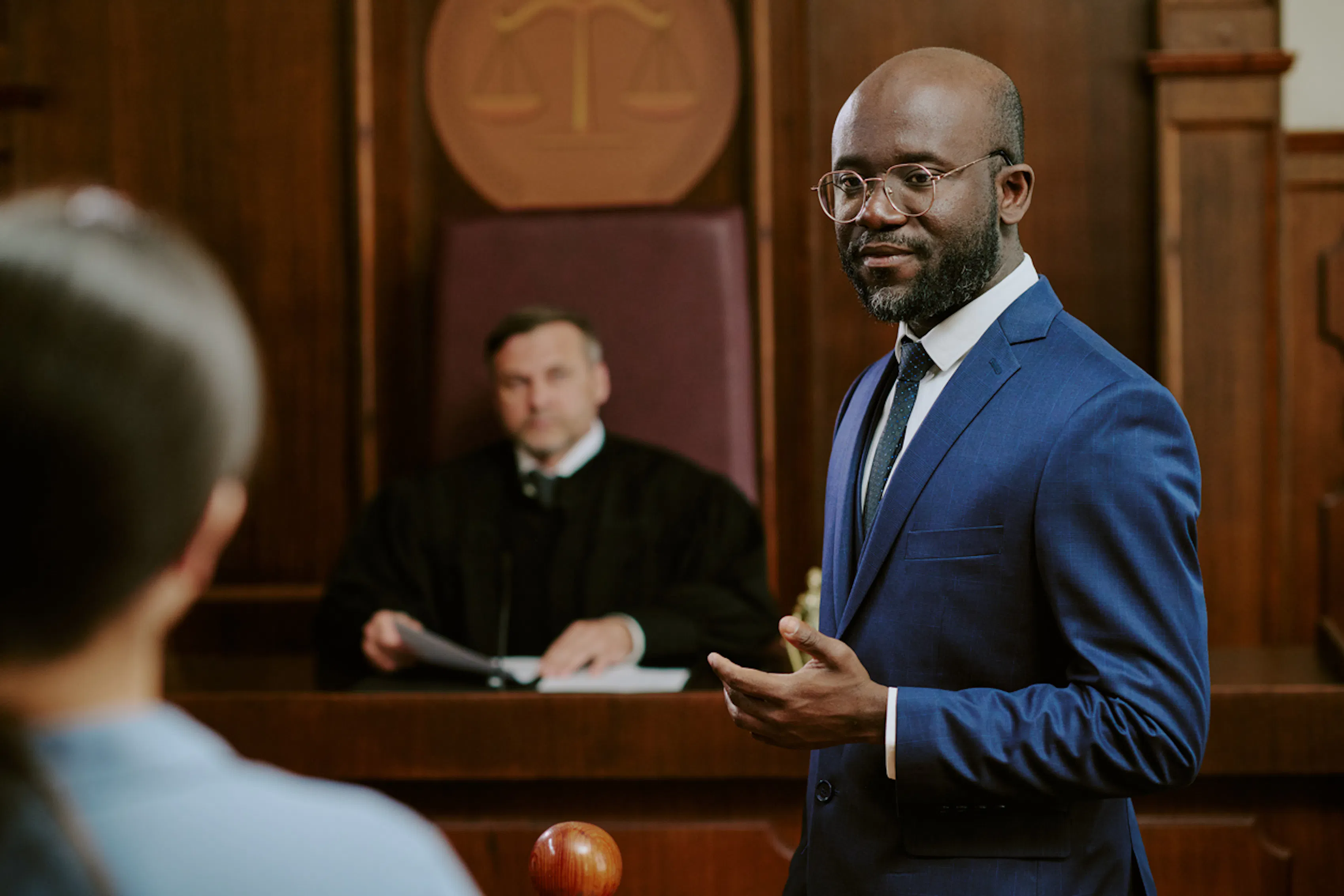 A man in a blue suit, wearing glasses, stands in a courtroom speaking. The judge, seated at the bench behind him, listens attentively. The courtroom is well-lit with wood paneling and a scale of justice emblem on the wall.