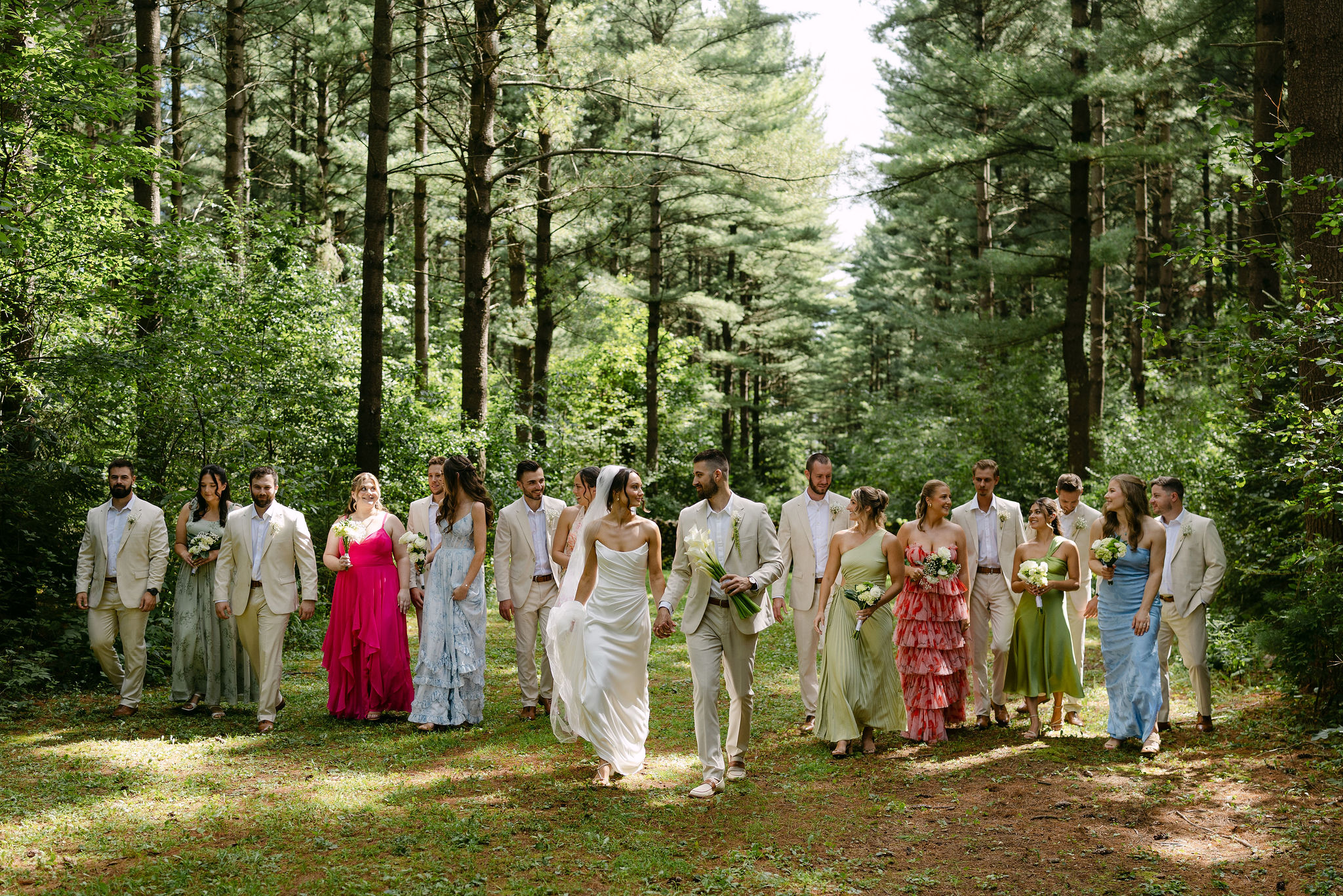 Bridal party walking through woods in colorful mix and match bridesmaid dresses and tan groomsmen suits