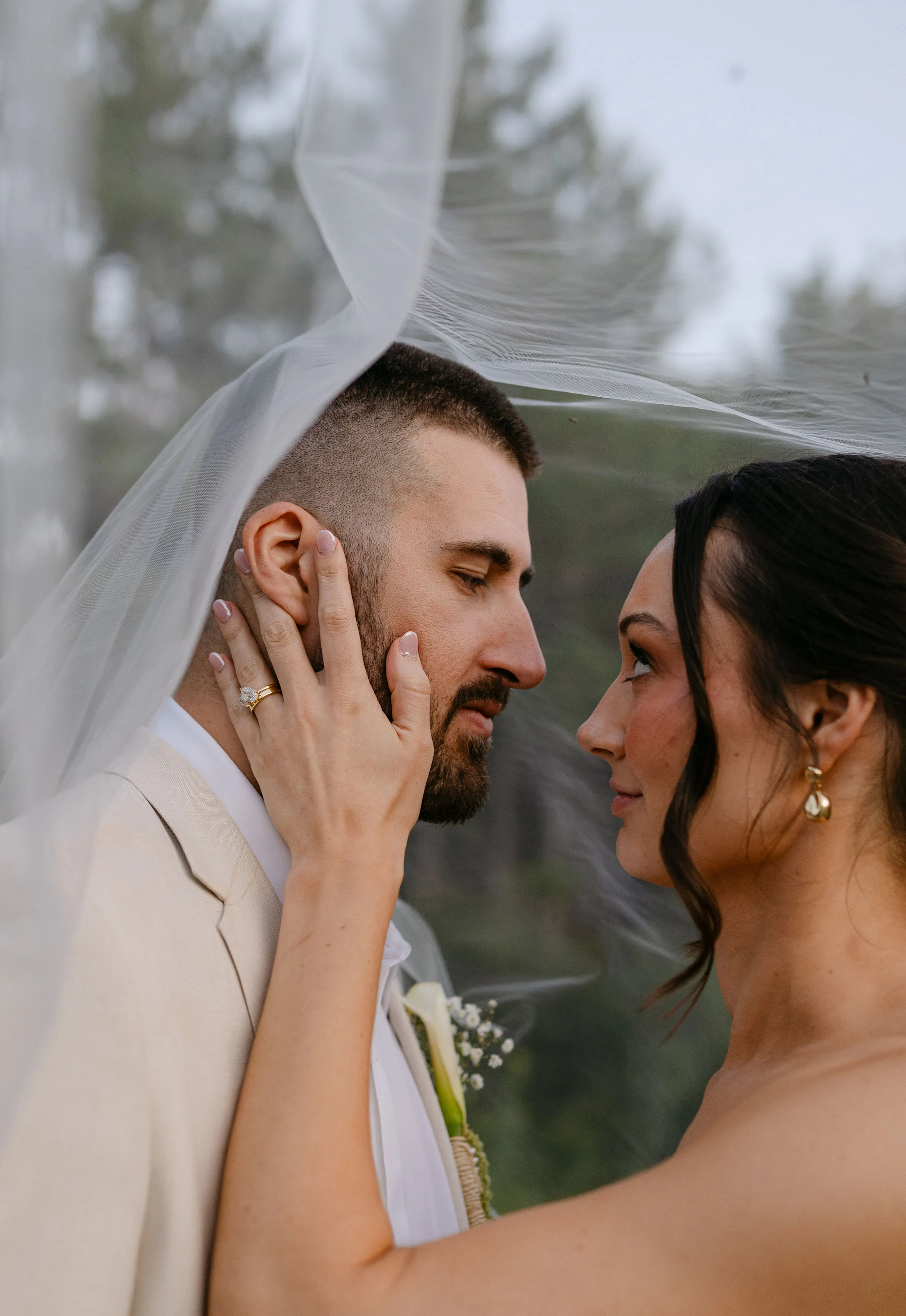 Bride with face framing hair style holding groom in tan wedding suit under veil