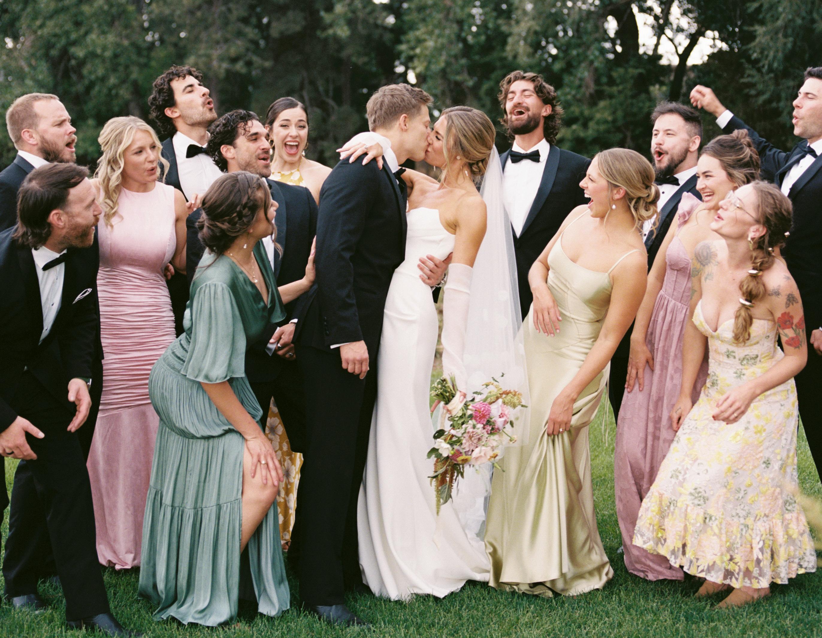 Wedding guests in summer dresses and tuxedos surrounding the newlywed bride and groom in a meadow.