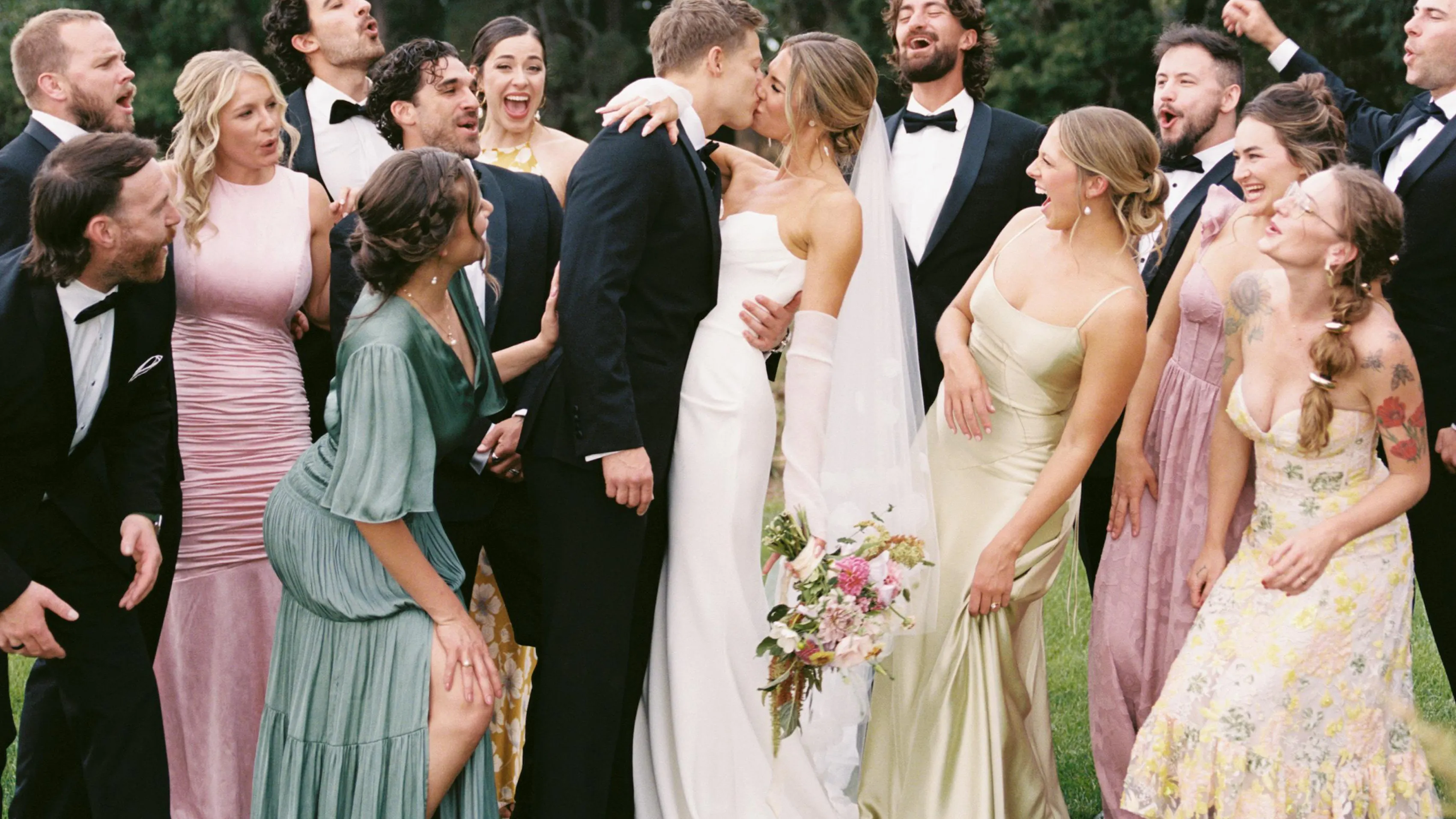 Wedding guests in summer dresses and tuxedos surrounding the newlywed bride and groom in a meadow.