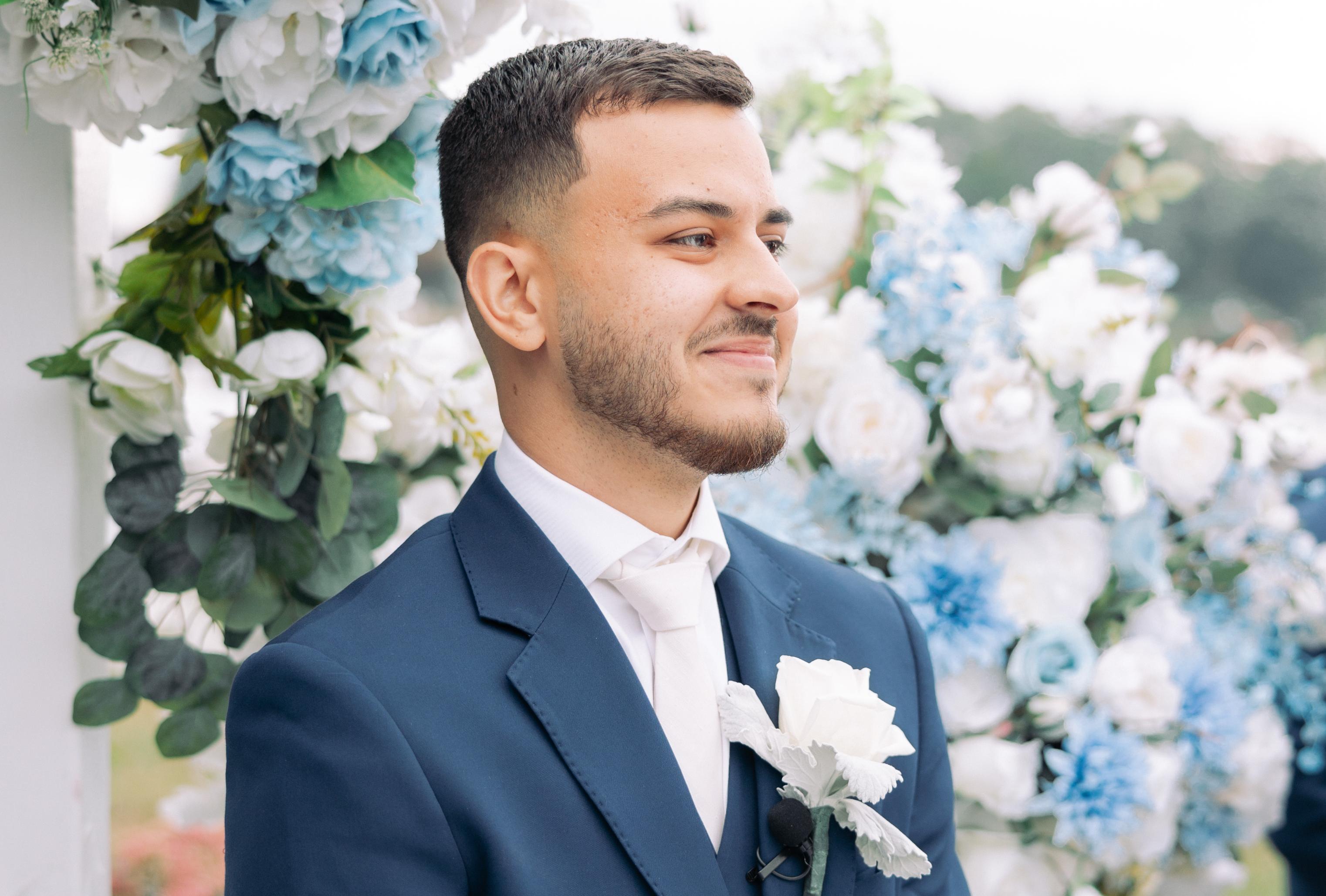 A man in a navy suit and white tie stands in front of a wall of blue and white roses, staring off into the distance.