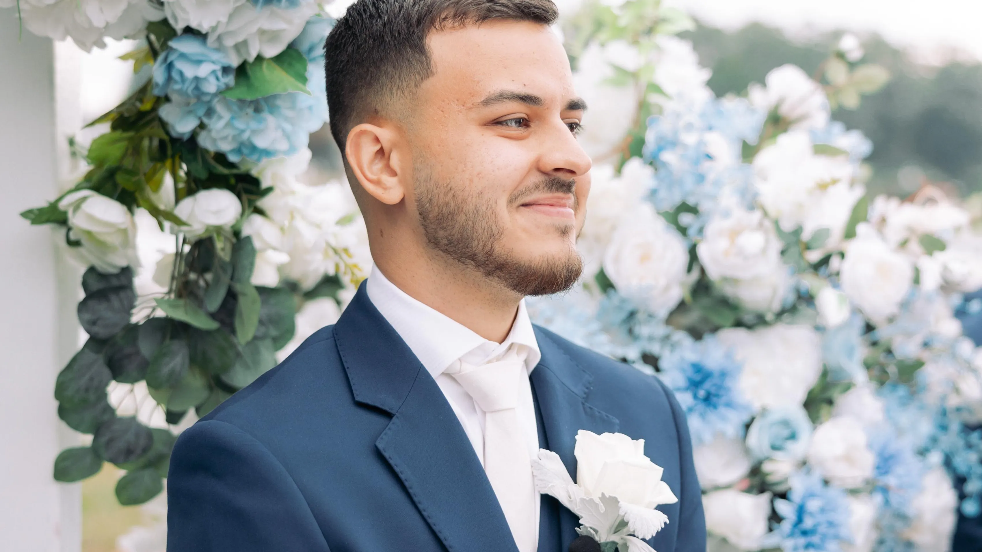A man in a navy suit and white tie stands in front of a wall of blue and white roses, staring off into the distance.