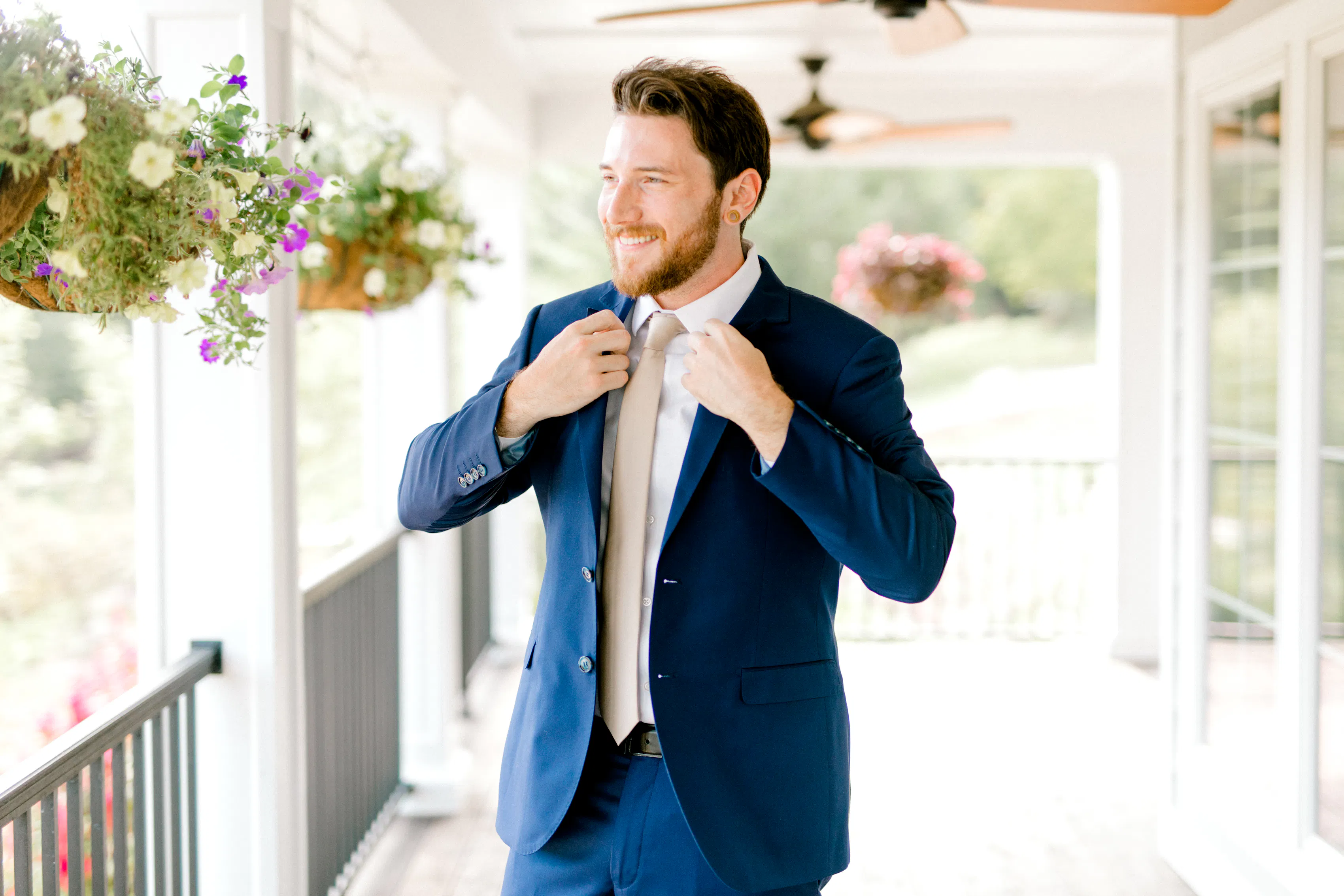 A man in a navy suit and beige tie adjusts his suit on a patio.