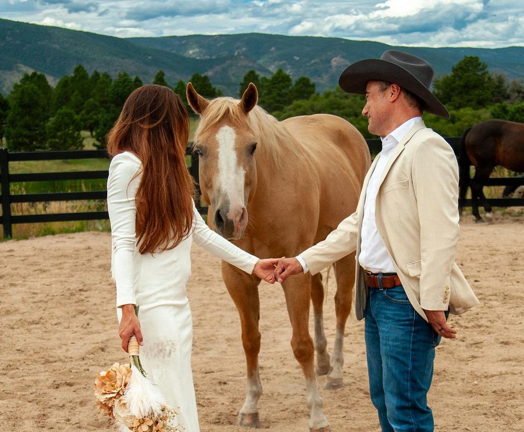 A man and woman at their wedding face eachother in front of a horse in a fenced in barn lot.