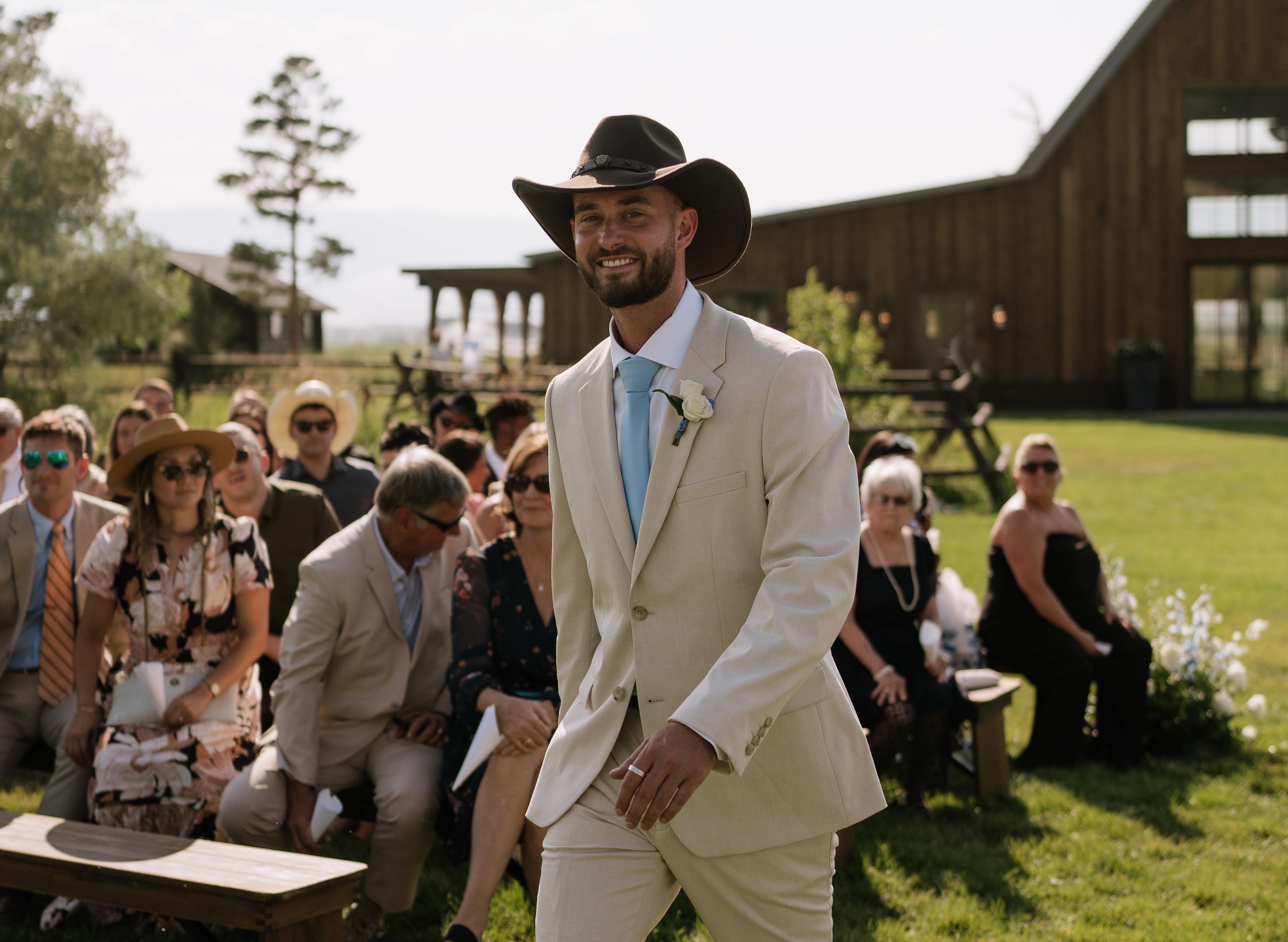 A groom walking down the aisle in a cowboy hat and tan suit at his barn wedding venue.
