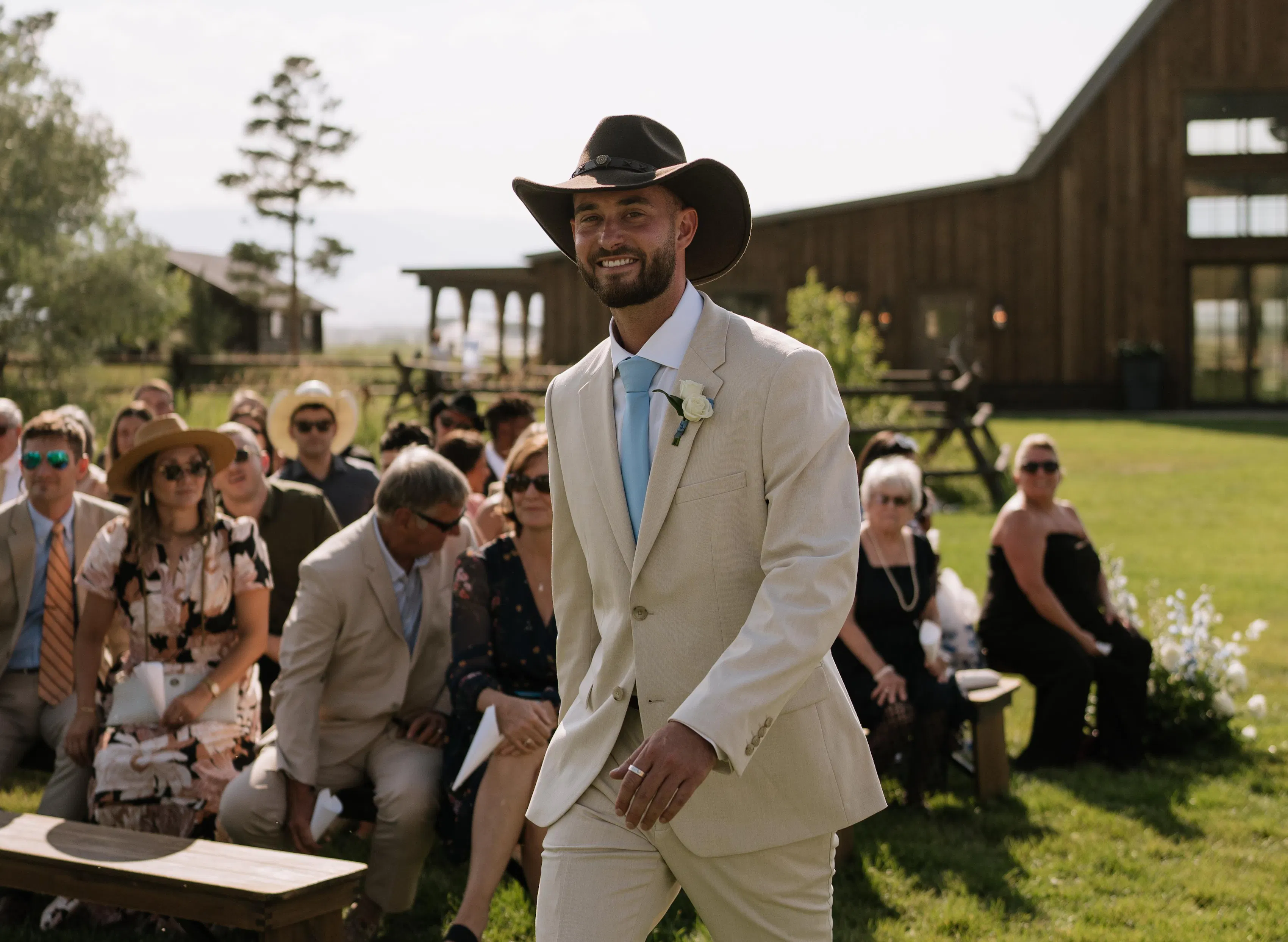 A groom walking down the aisle in a cowboy hat and tan suit at his barn wedding venue.