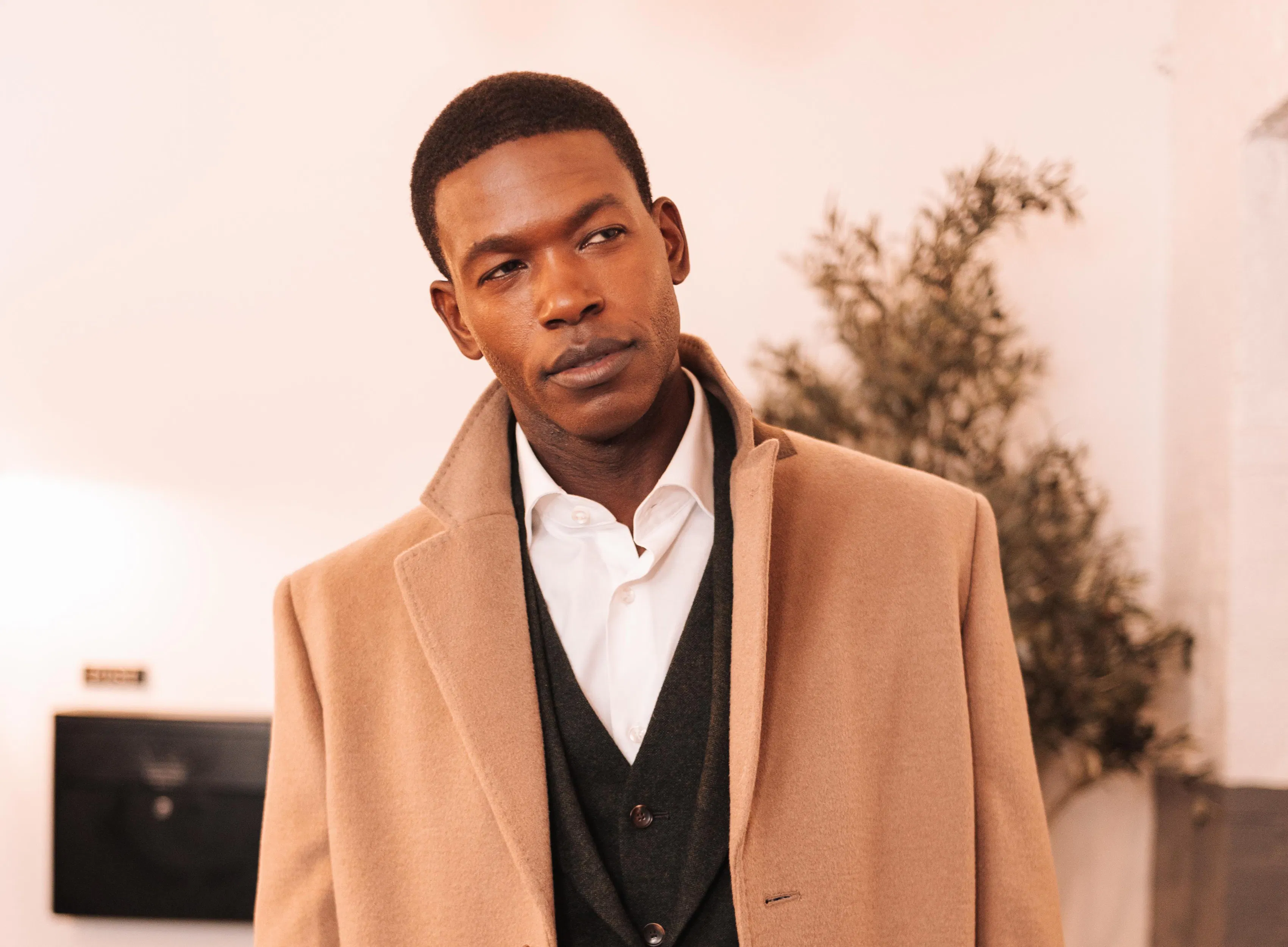 A man in a 3 piece suit under a camel trenchcoat looks to the side.