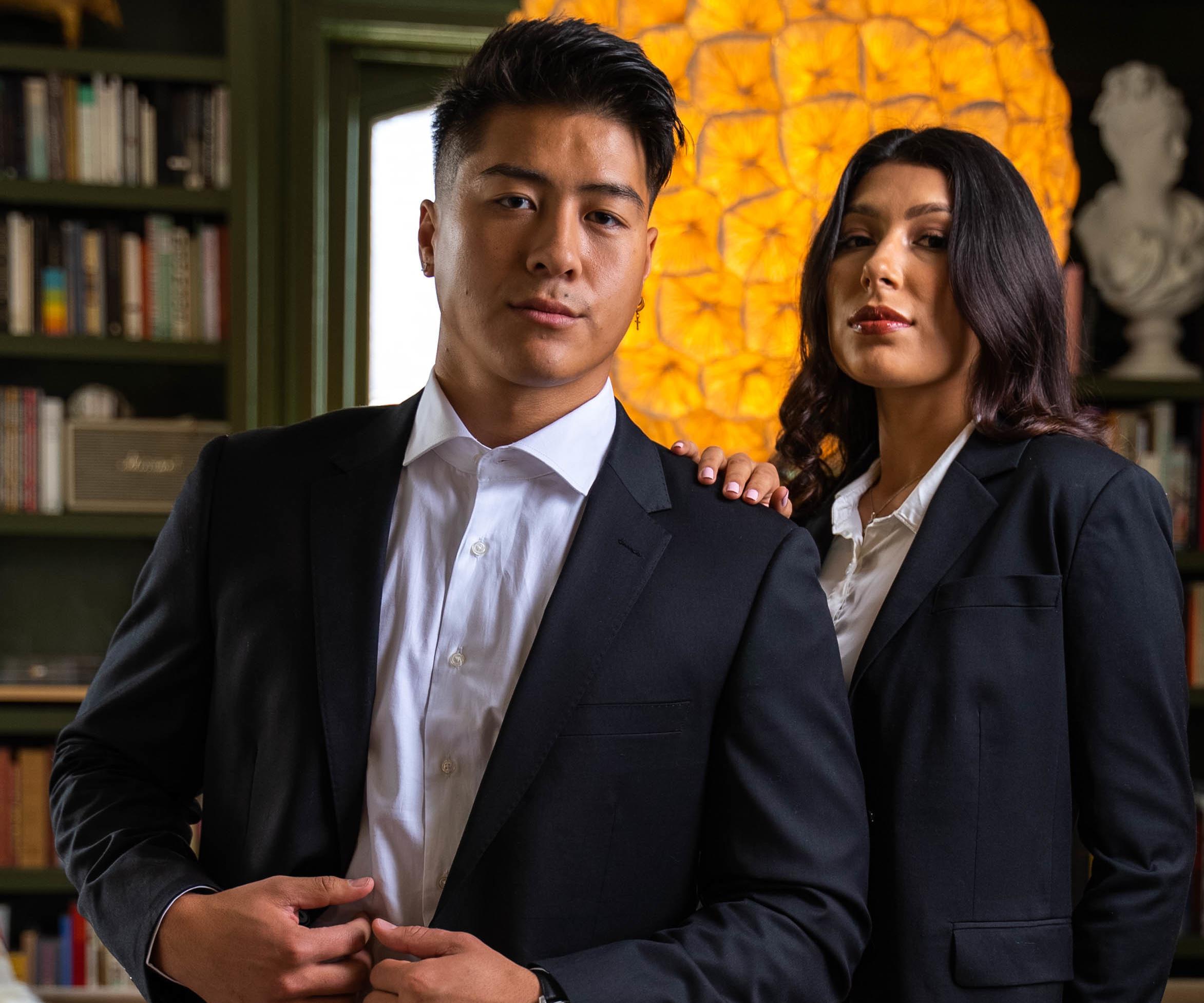 A man and woman stand in an office den wearing suits, posed confidently.