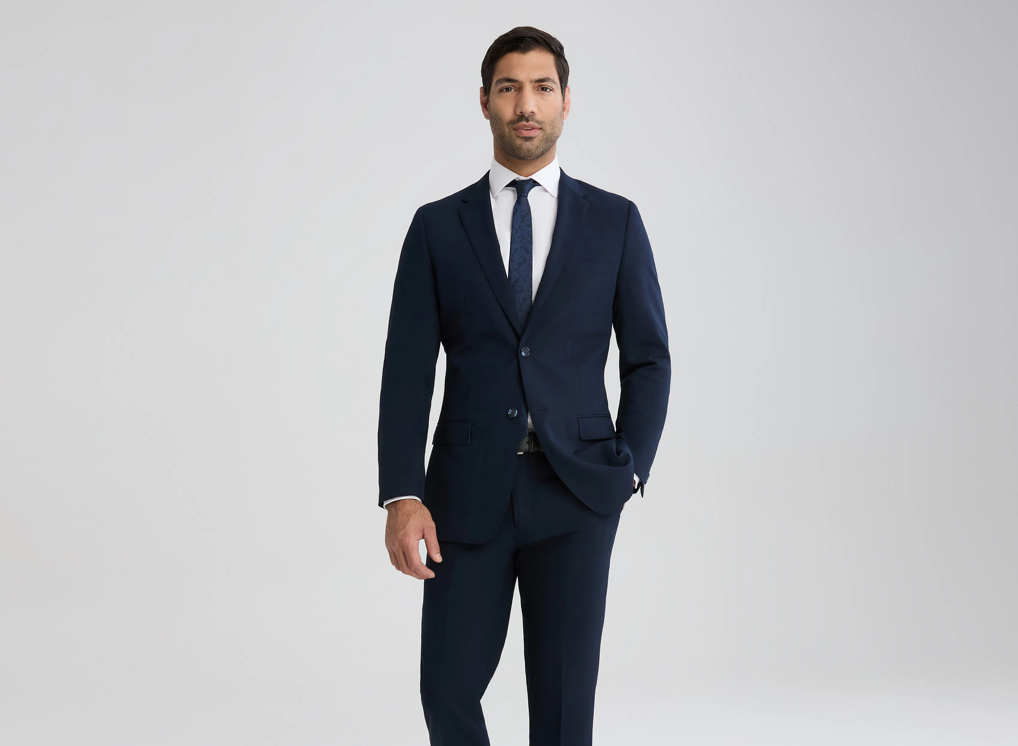 A man in a white dress shirt, blue tie, and navy blue suit stands in front of a blank background with his hand in his pocket.