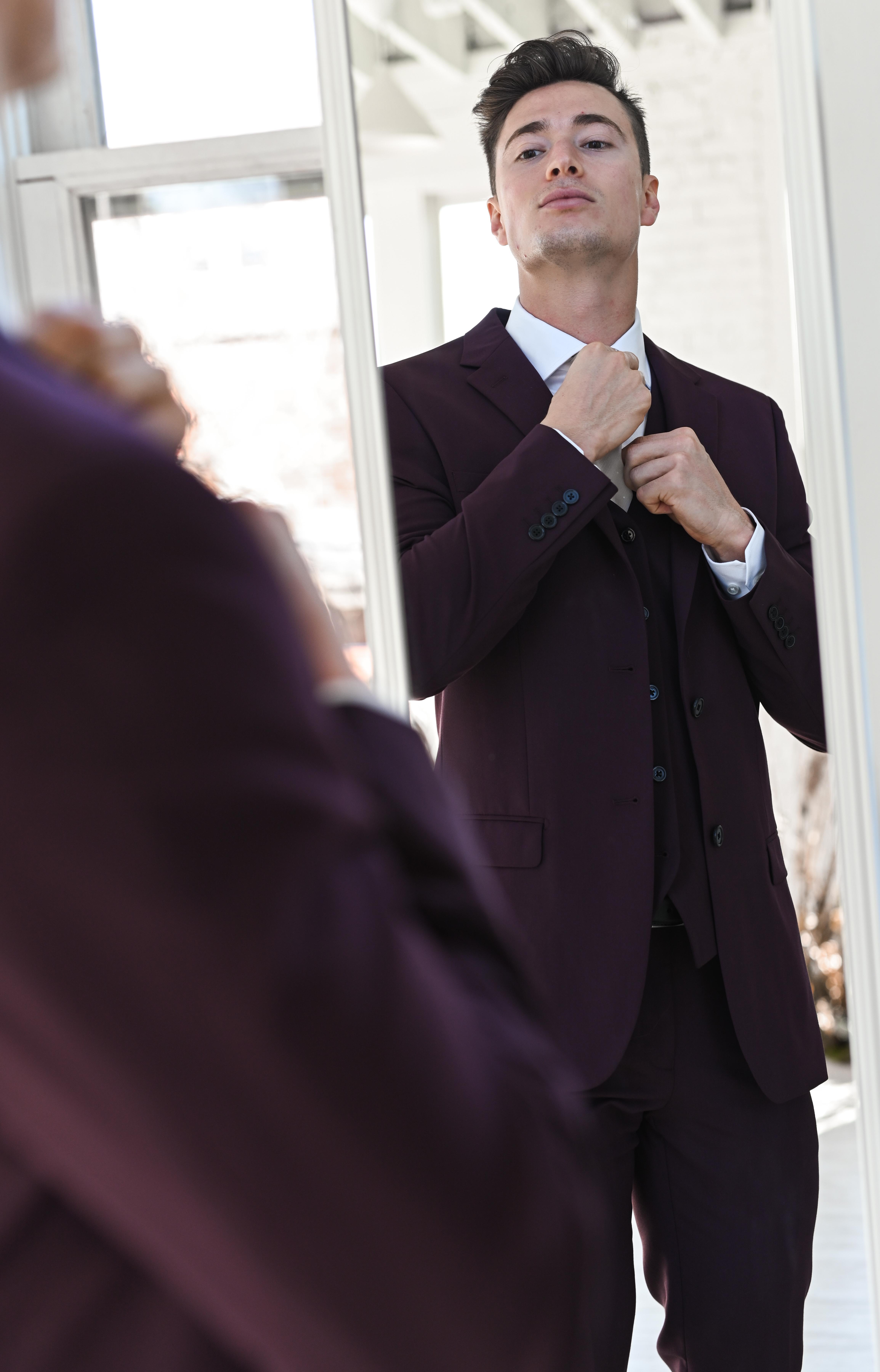 A man in a maroon suit adjusts his tie in the mirror, preparing for a wedding.