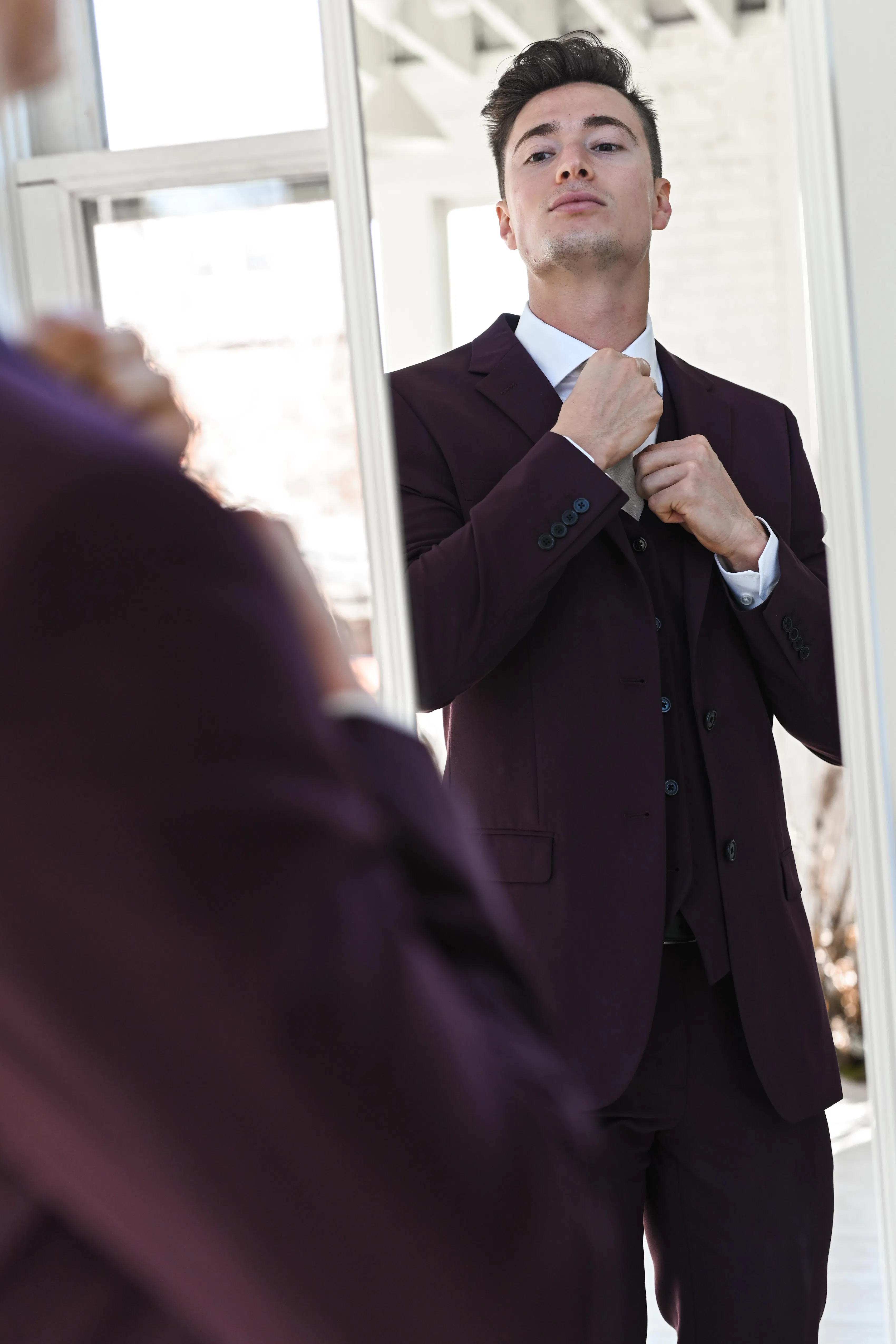 A man in a maroon suit adjusts his tie in the mirror, preparing for a wedding.