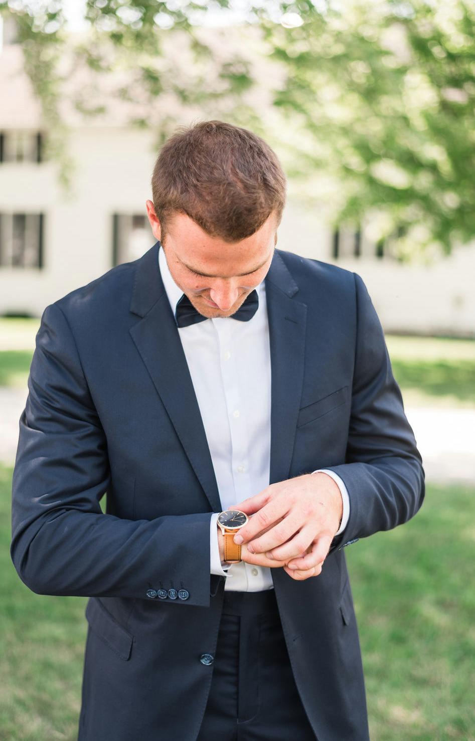 A man in a tuxedo adjusts his watch in an outdoor wedding venue.