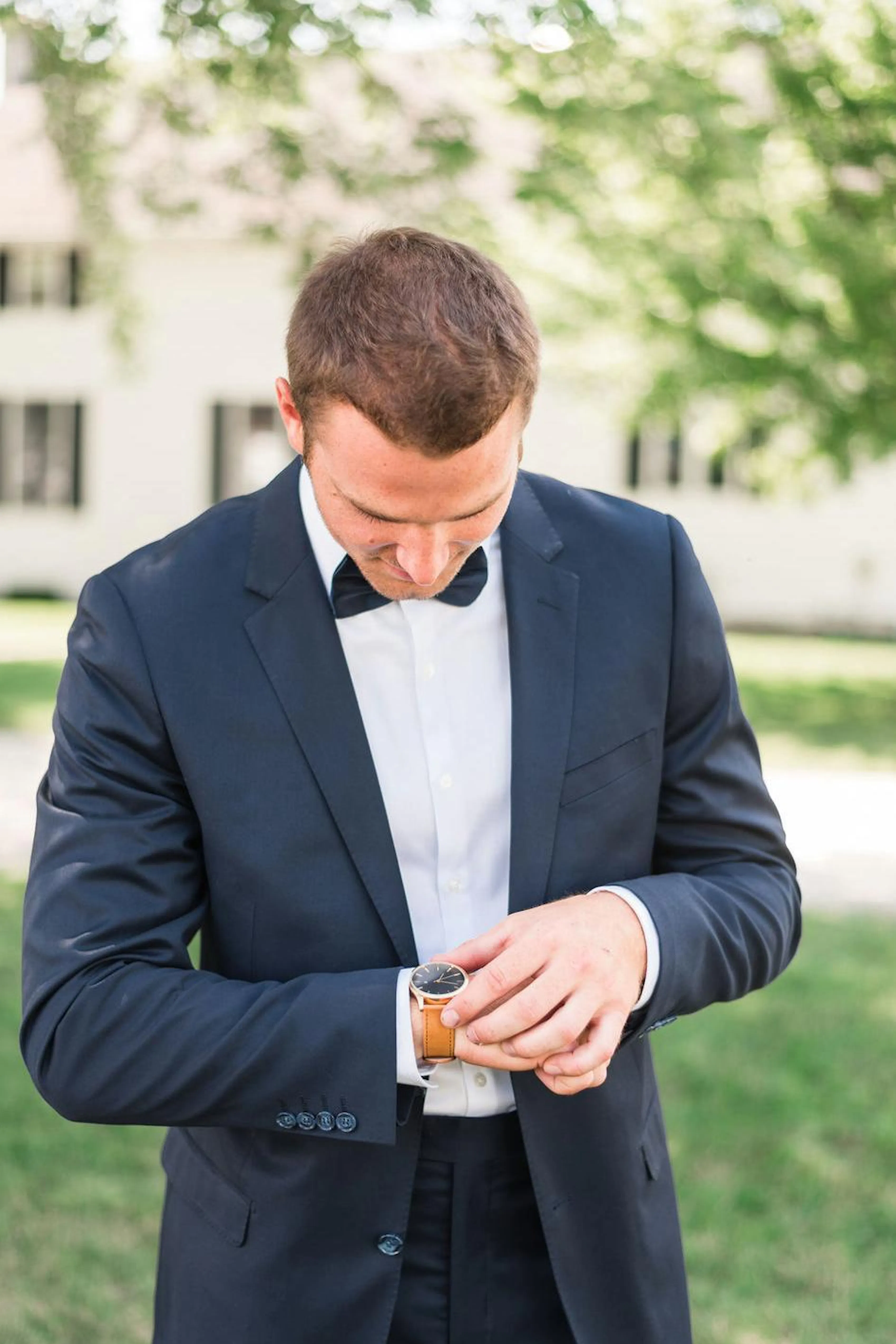 A man in a tuxedo adjusts his watch in an outdoor wedding venue.
