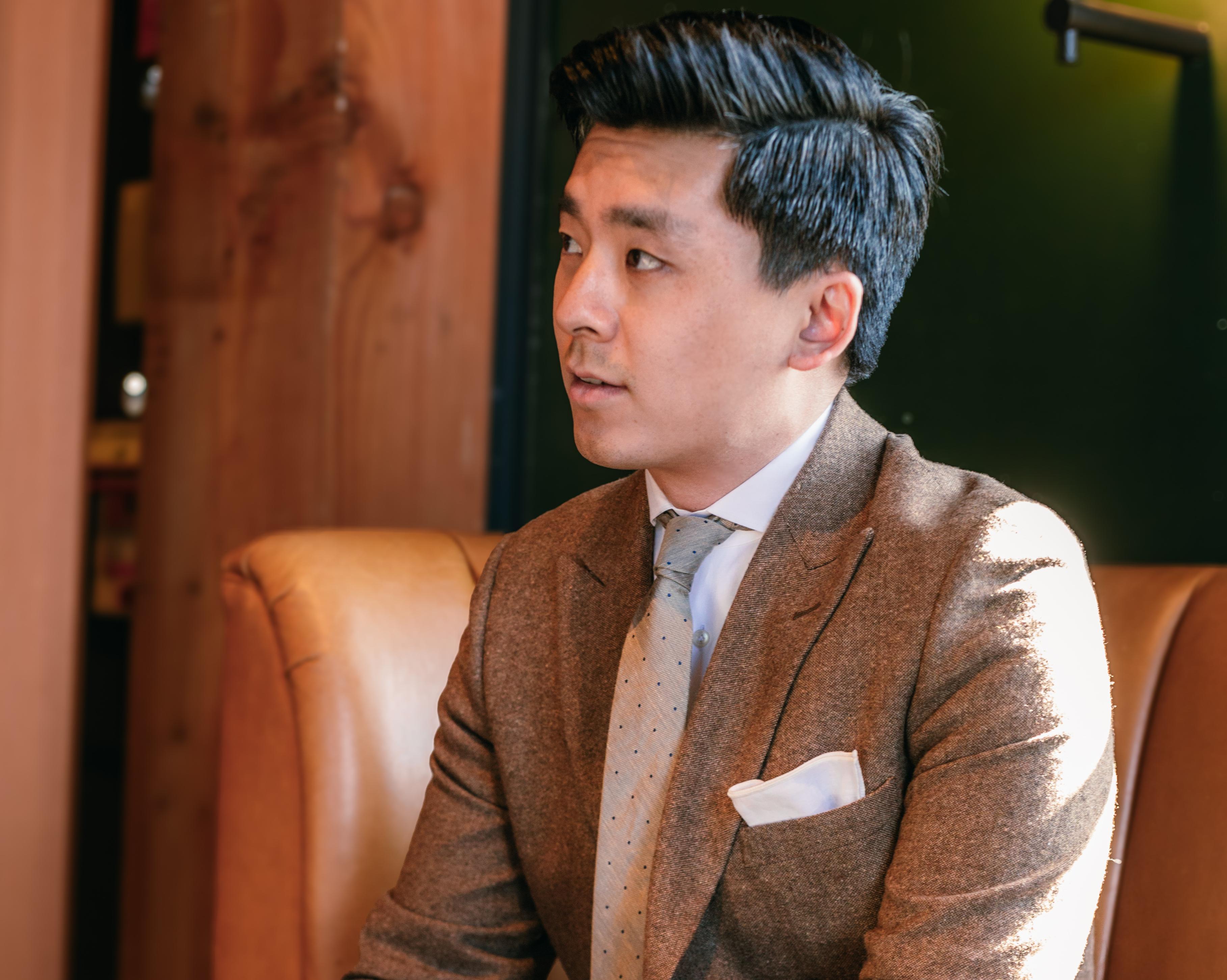 A man in cocktail attire, specifically a brown tweed suit with a tan tie, sits on a mid-century modern couch and stares into the distance.