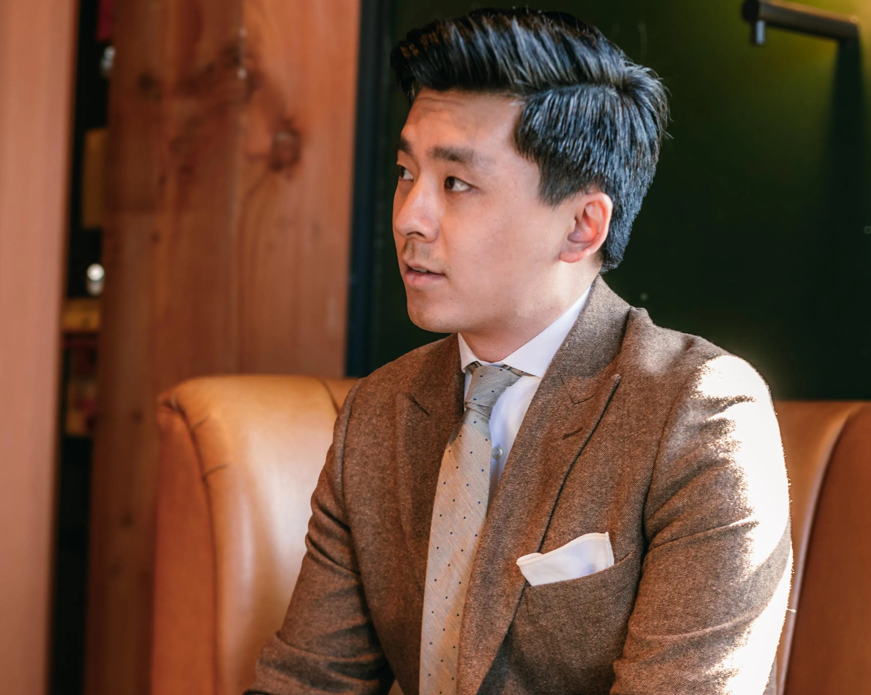 A man in cocktail attire, specifically a brown tweed suit with a tan tie, sits on a mid-century modern couch and stares into the distance.