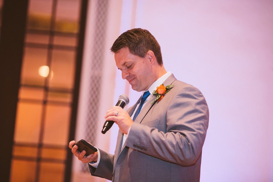 Man in light grey suit at wedding reception reading a speech to honor the loss of a parent.