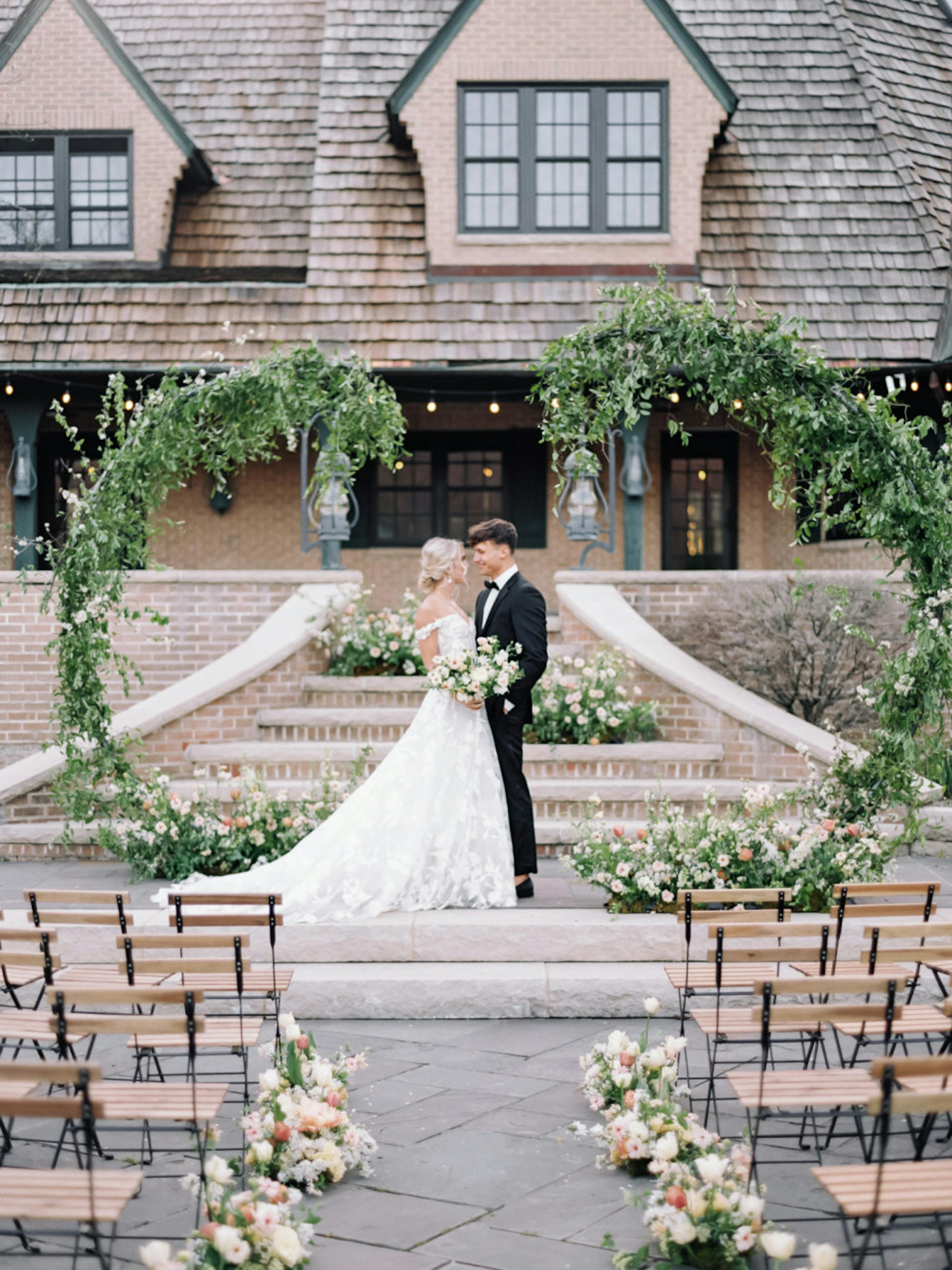 Bride in A-line lace wedding gown and groom wearing classic wedding tuxedo standing at the altar under green ivy arches.