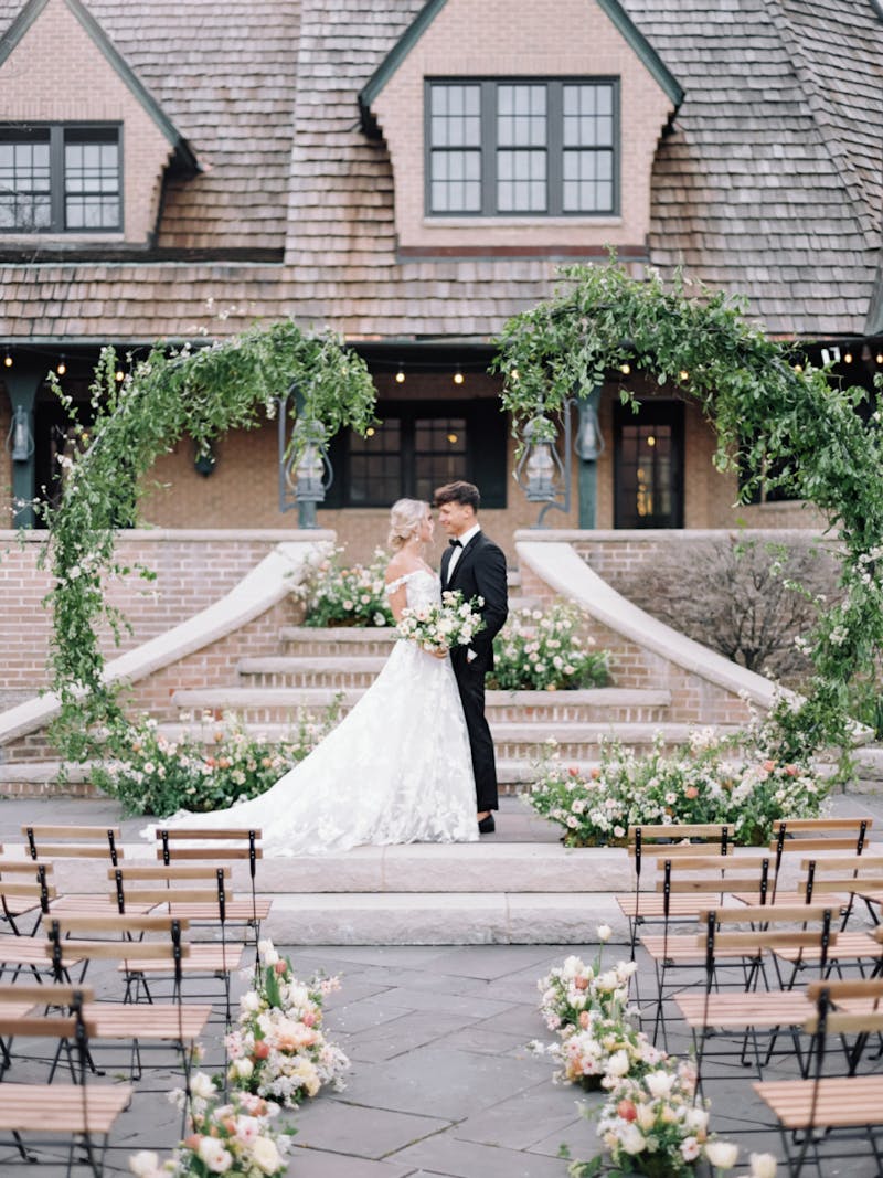 Bride in A-line lace wedding gown and groom wearing classic wedding tuxedo standing at the altar under green ivy arches.