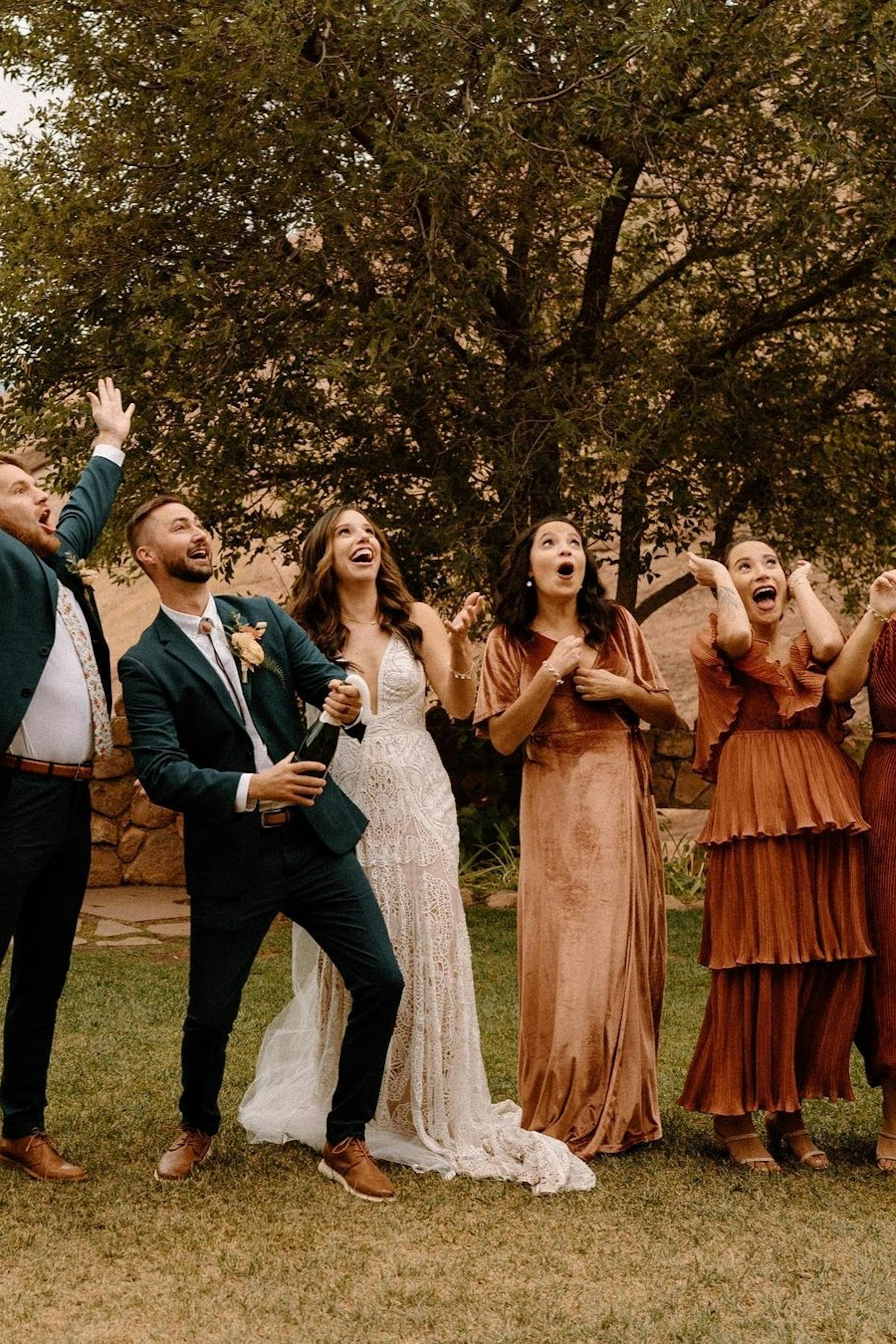 Western wedding party popping champagne with groom in dark green suit and bolo tie and rust bridesmaids dresses.