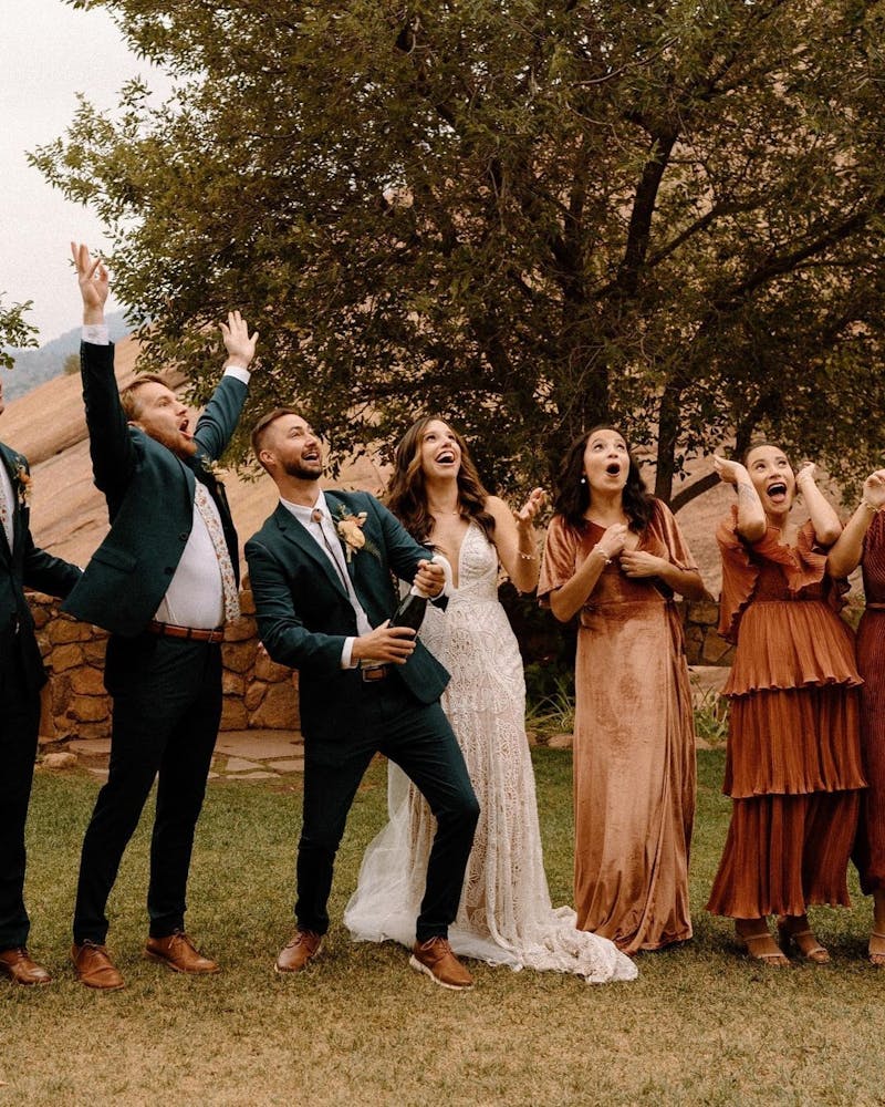 Western wedding party popping champagne with groom in dark green suit and bolo tie and rust bridesmaids dresses.