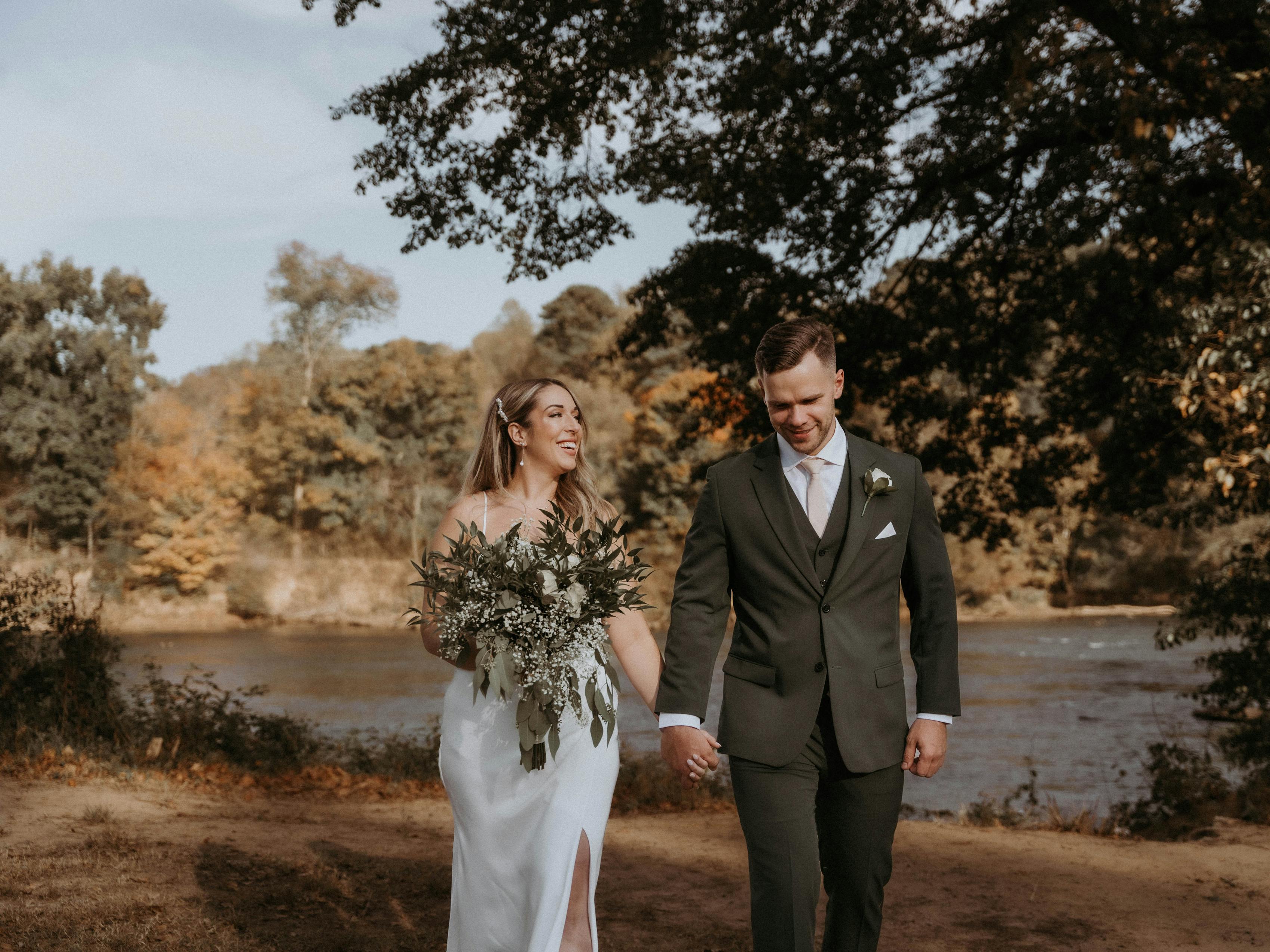 Dark green suit and vest on groom walking with modern bride at lakeside wedding.