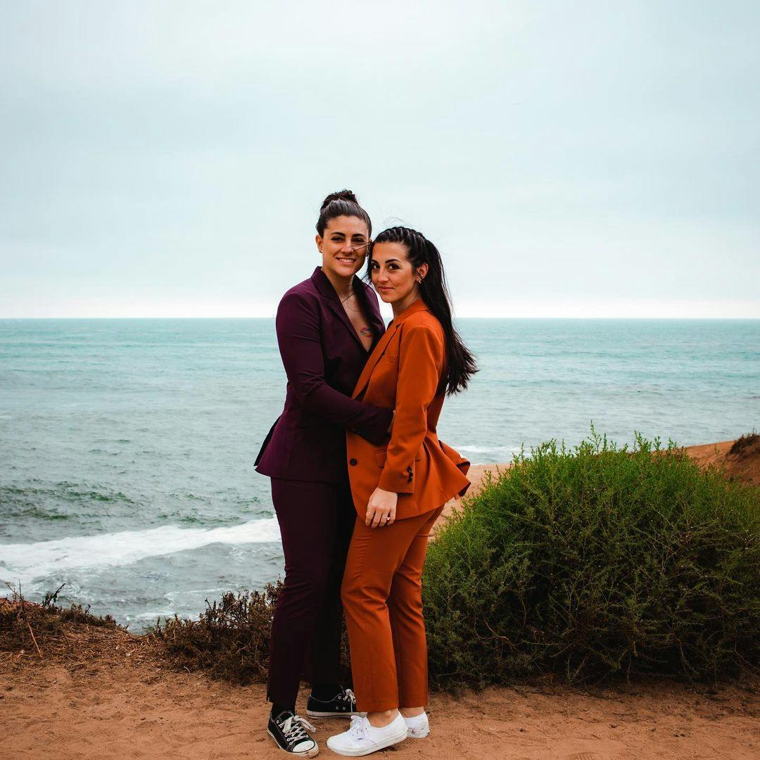 Lesbian couple style in women's maroon burgundy suit and women's burnt orange suit paired with sneakers on the beach.