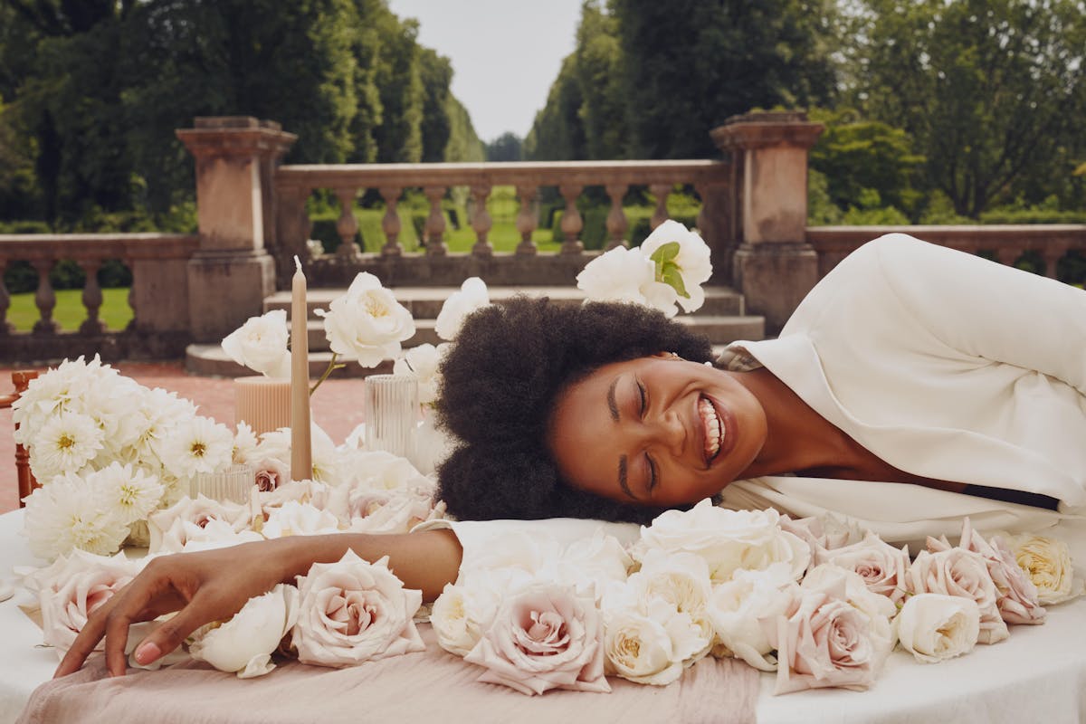 Eyes closed in moment of joy, blushing bride wears a white tuxedo dress and lies on a wedding tablescape covered in light colored wedding flowers.