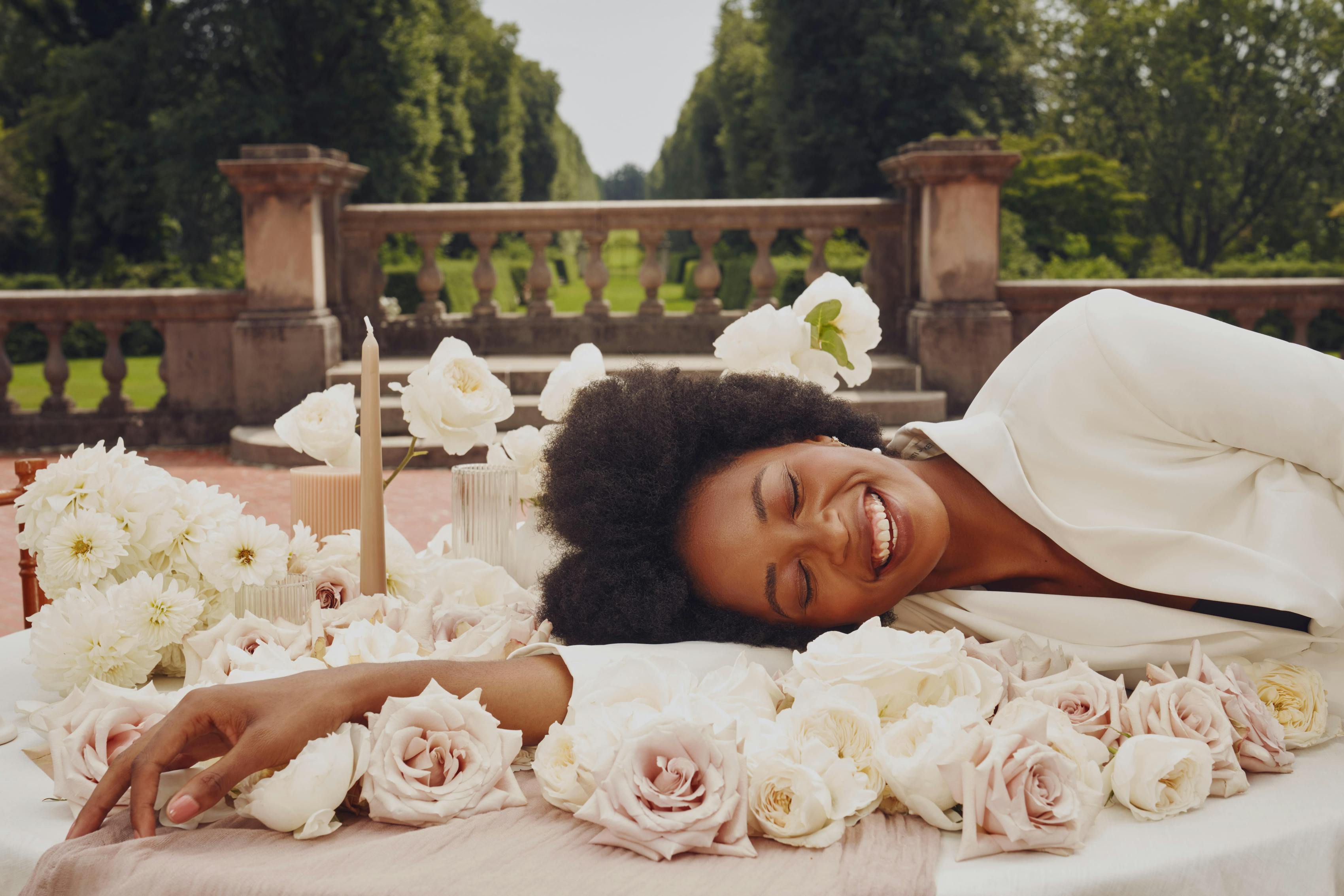 Eyes closed in moment of joy, blushing bride wears a white tuxedo dress and lies on a wedding tablescape covered in light colored wedding flowers.