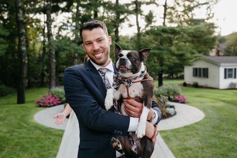 Man in navy blue suit and dark floral neck tie holding a dog at a summer wedding.