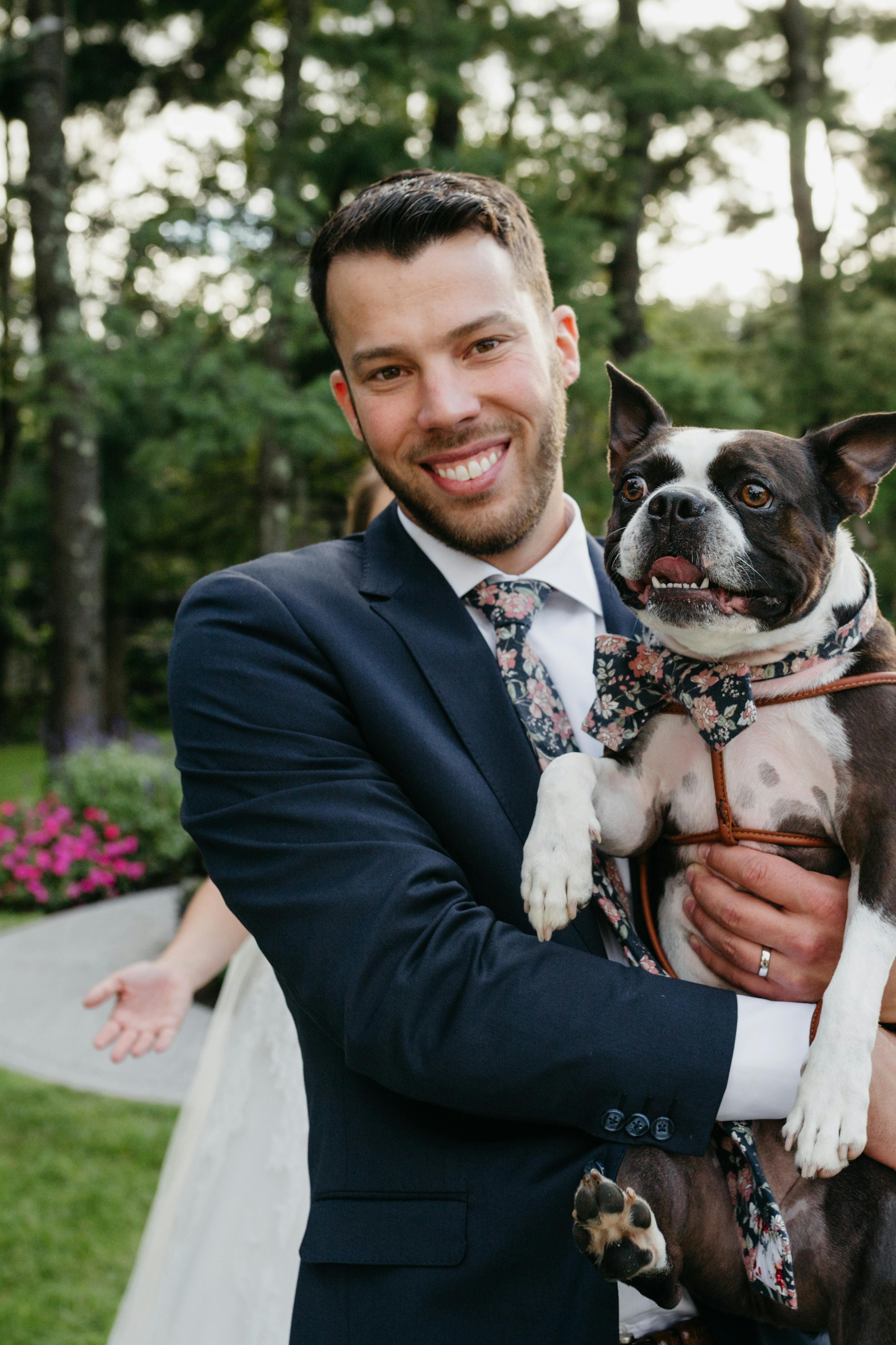 Man in navy blue suit and dark floral neck tie holding a dog at a summer wedding.