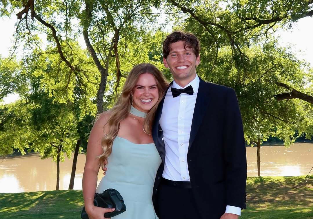 Couple posing outdoors by a lake for a fraternity formal, with the man wearing a black tuxedo and bow tie and the woman in a light blue strapless dress.