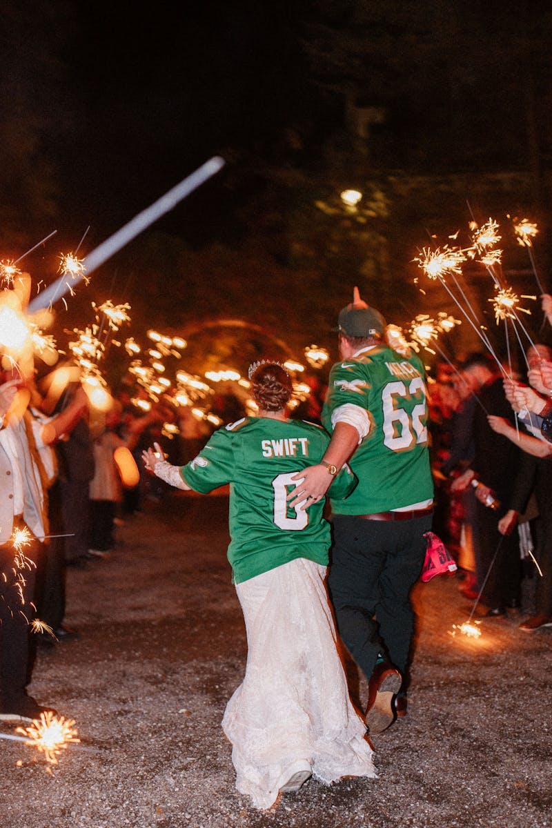 Football themed wedding exit with bride and groom wearing Eagles jerseys under a sparkler arch.