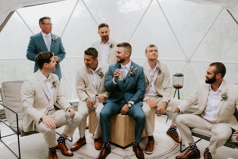 Outdoorsy groomsmen group drinking beer in tan groomsmen suits and light blue groom suit and father of the groom suit.