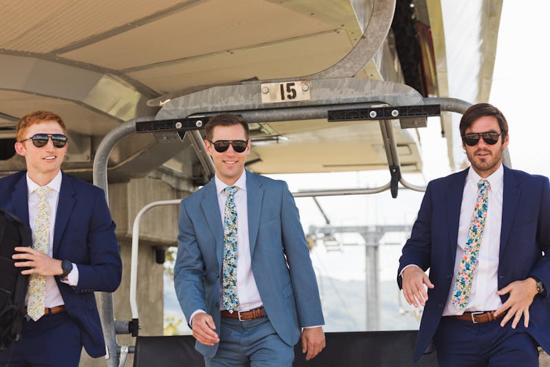 Three men at a mountain wedding wearing blue suits, floral ties, and sunglasses on a chairlift.
