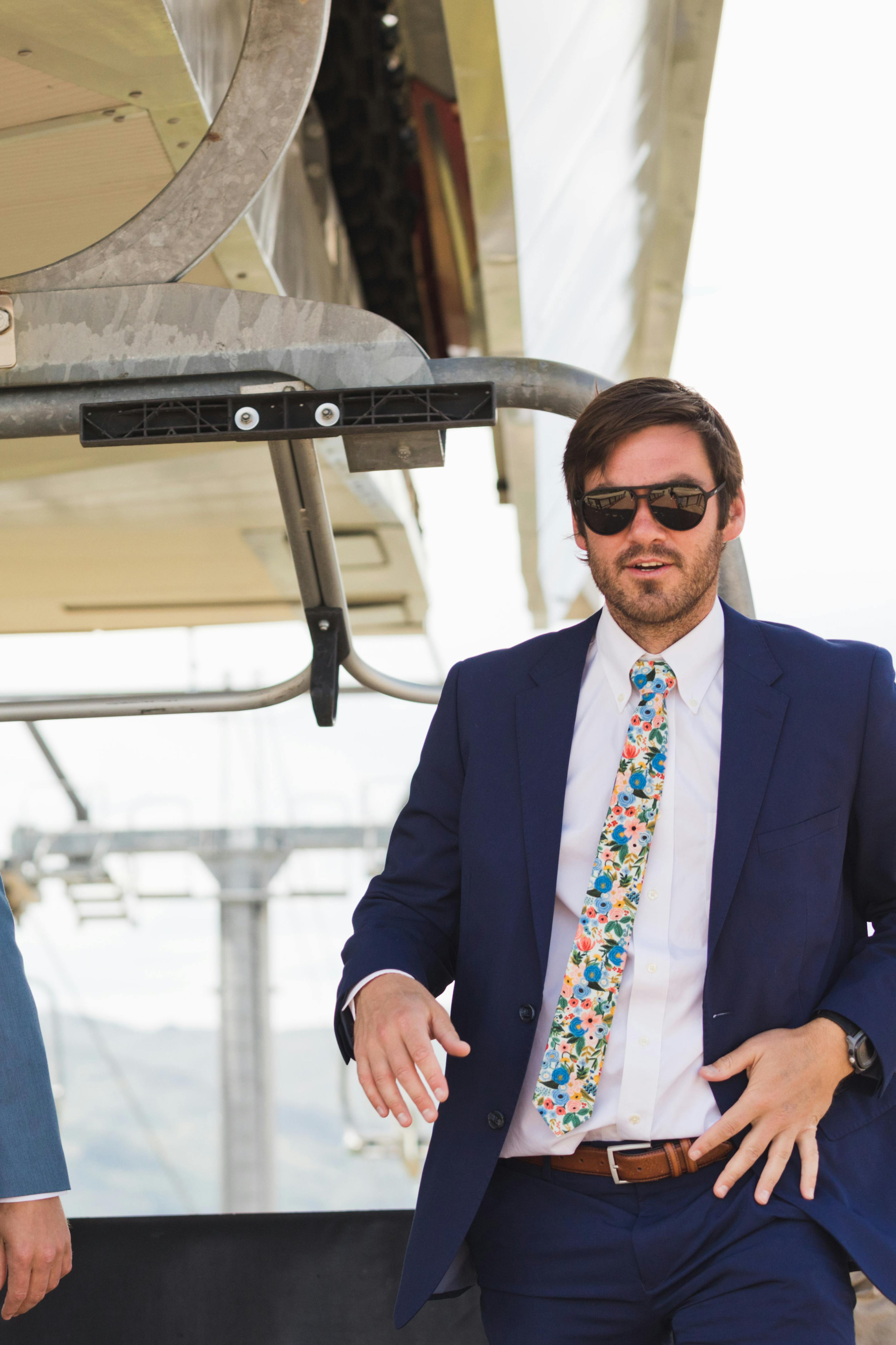 Three men at a mountain wedding wearing blue suits, floral ties, and sunglasses on a chairlift.