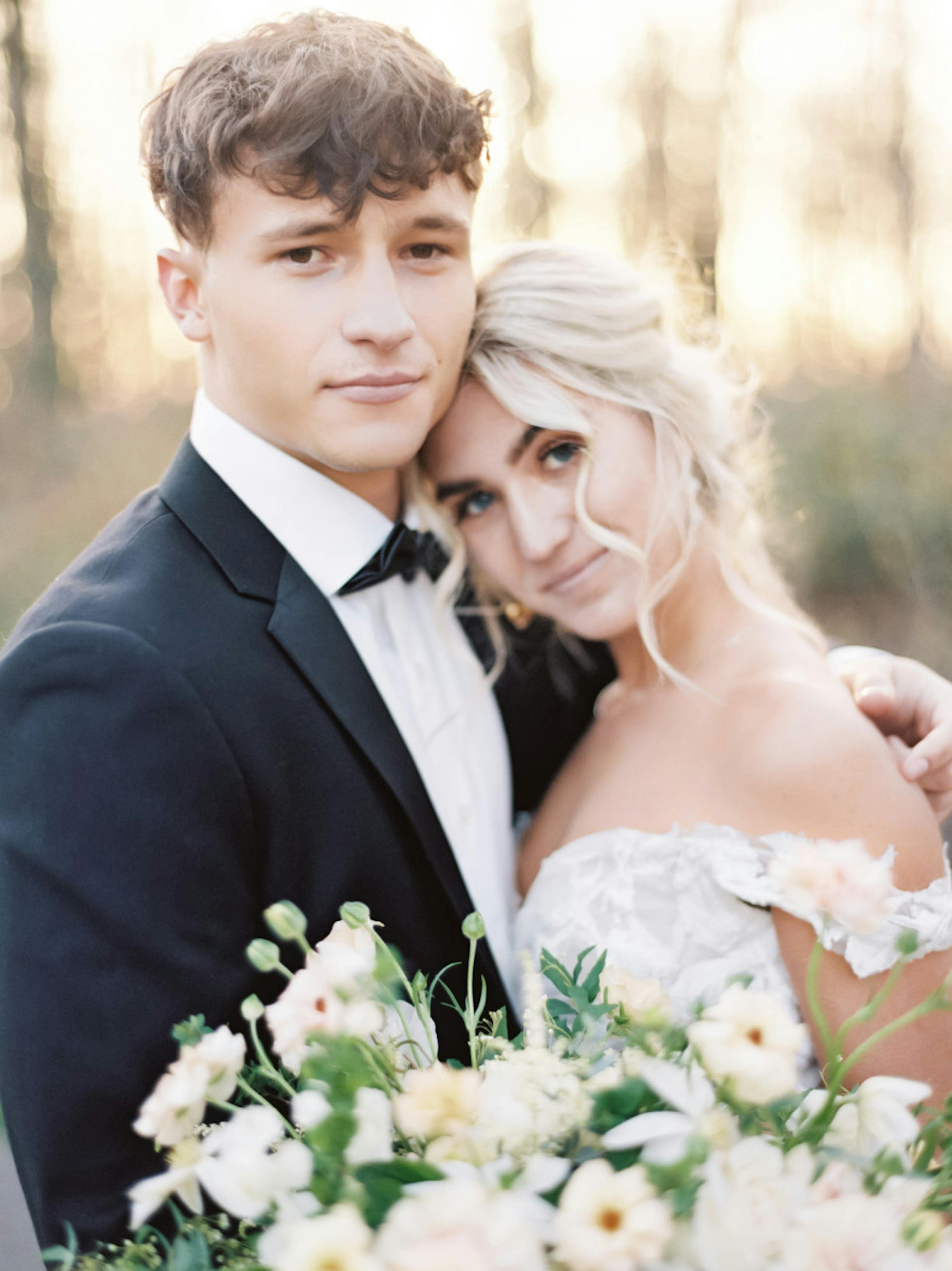 Groom wearing notch lapel black tuxedo and bow tie hugging bride wearing off the shoulder wedding dress. 